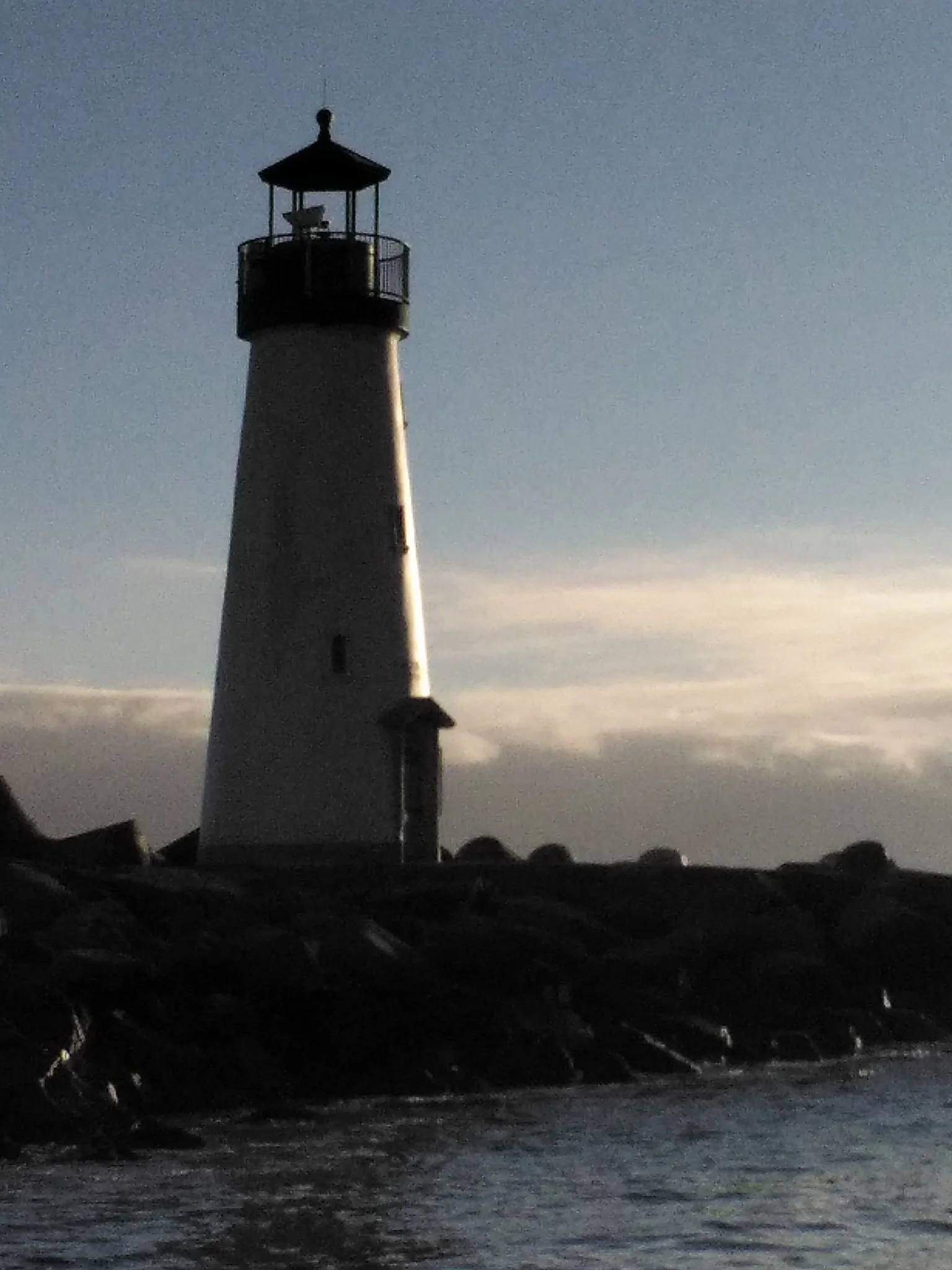 Santa Cruz Breakwater (aka Walton) Lighthouse, located on the Santa Cruz Harbor, standing as a beacon during dusk with the sunlight reflecting off of it and a partly cloudy sky.