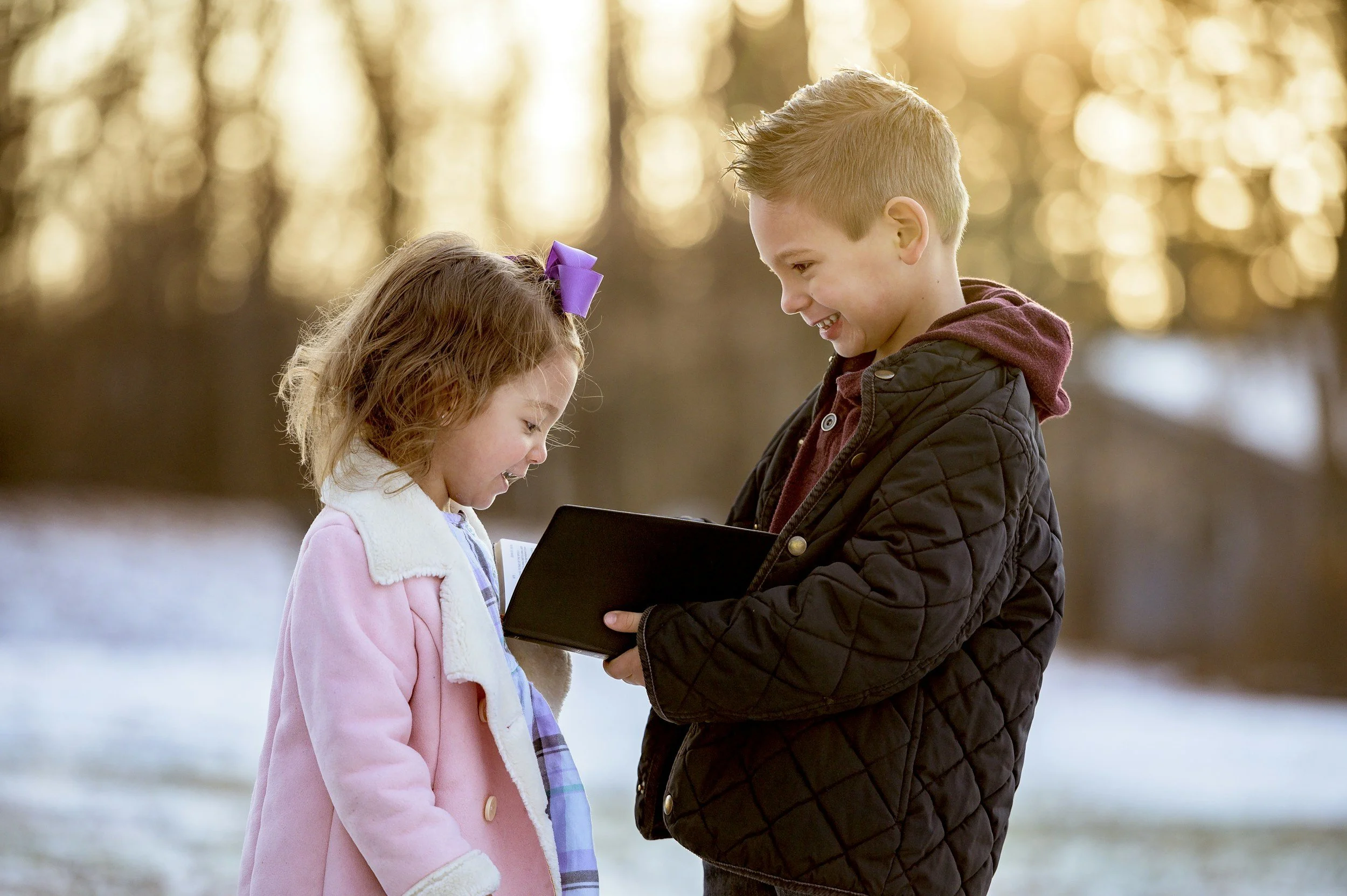 A boy and girl outdoors in winter, smiling and looking at a phone or small device together.