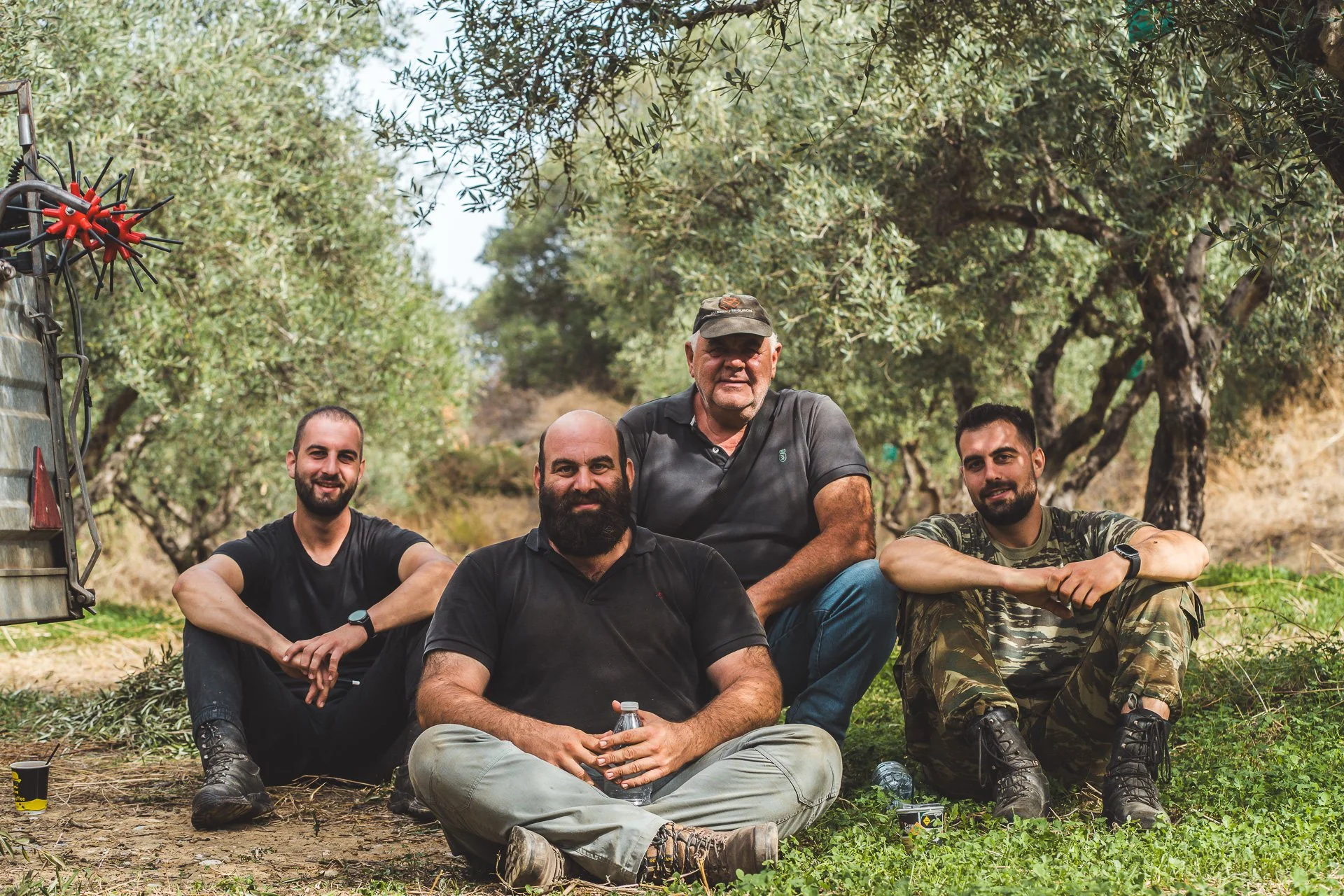 Four men sitting outdoors on the ground in a natural setting with trees, posing for a photo, with equipment visible on the left side.