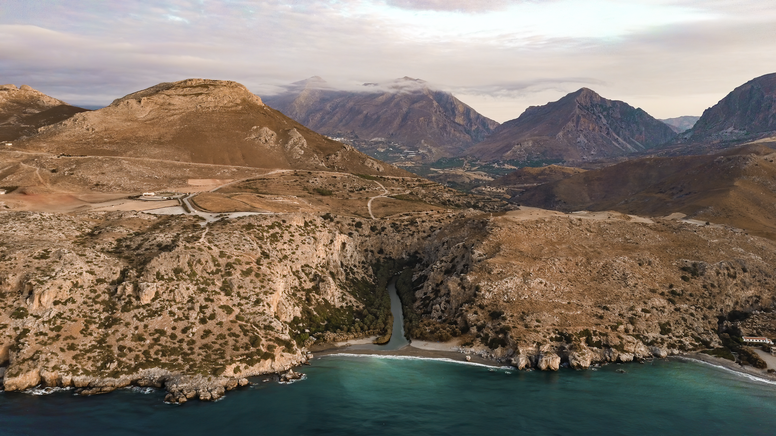 Scenic view of a rugged coastline with cliffs and a narrow river flowing into the ocean, surrounded by brown, arid hills and mountains under a cloudy sky.