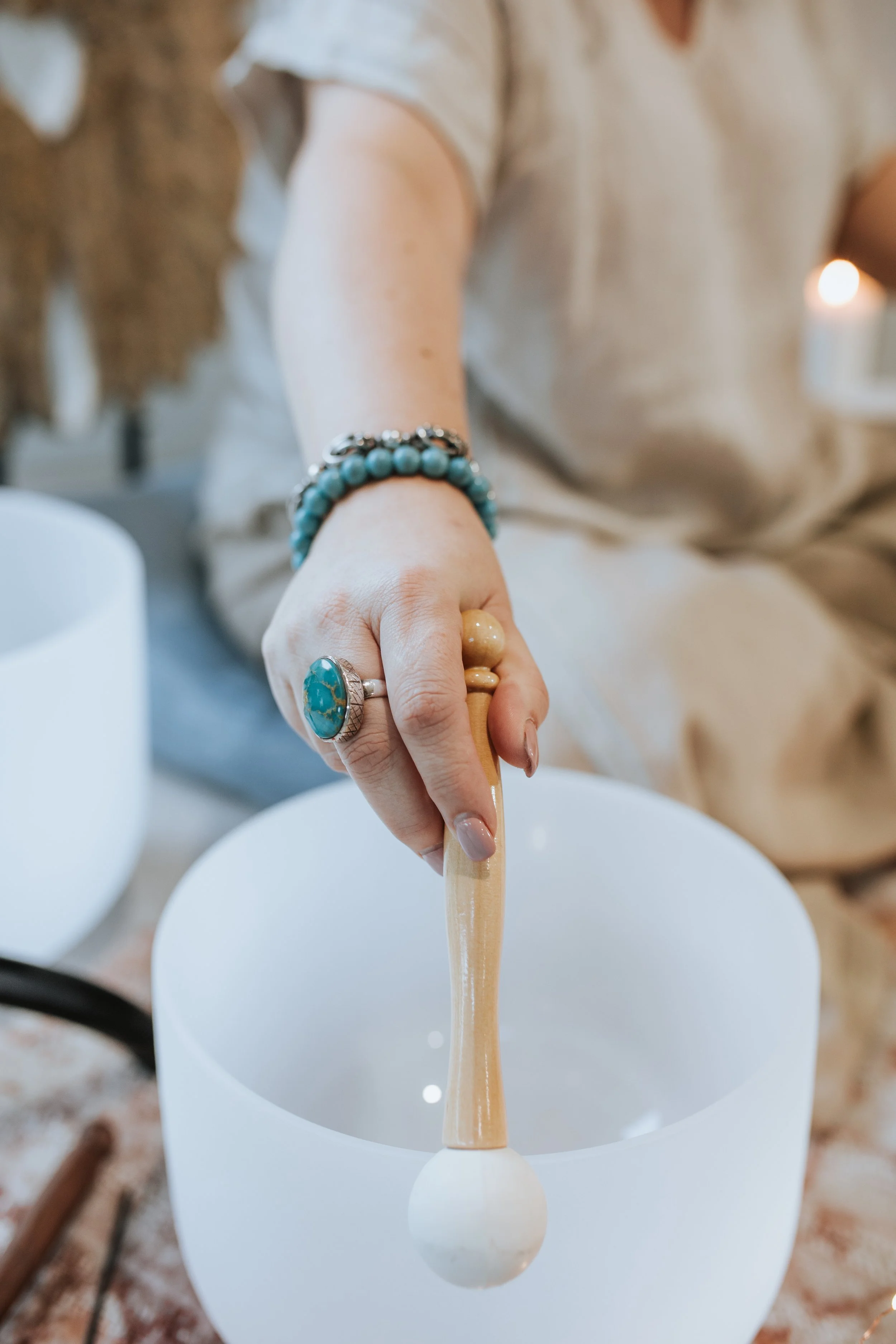 A person holding a mallet over a white crystal singing bowl, with jewelry including rings and bracelets on their hand.