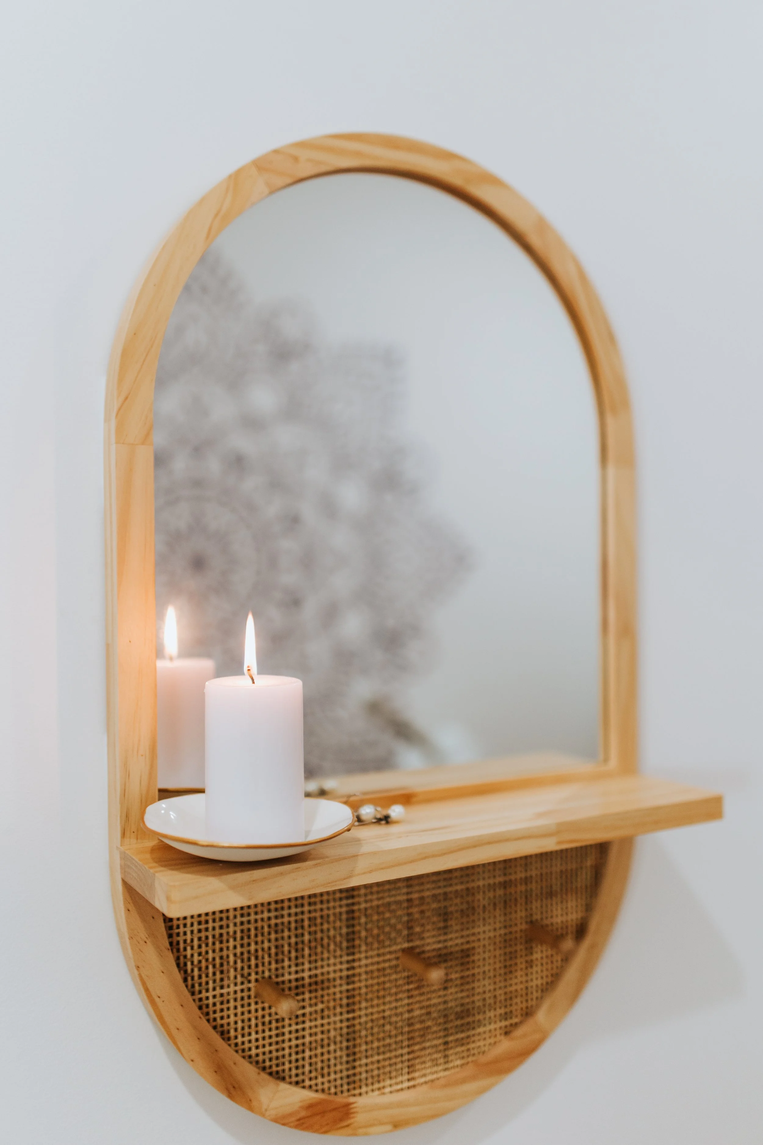 A round wooden-framed mirror with a small shelf holding a lit white candle in a dish, reflecting a blurred plant or decoration behind it.
