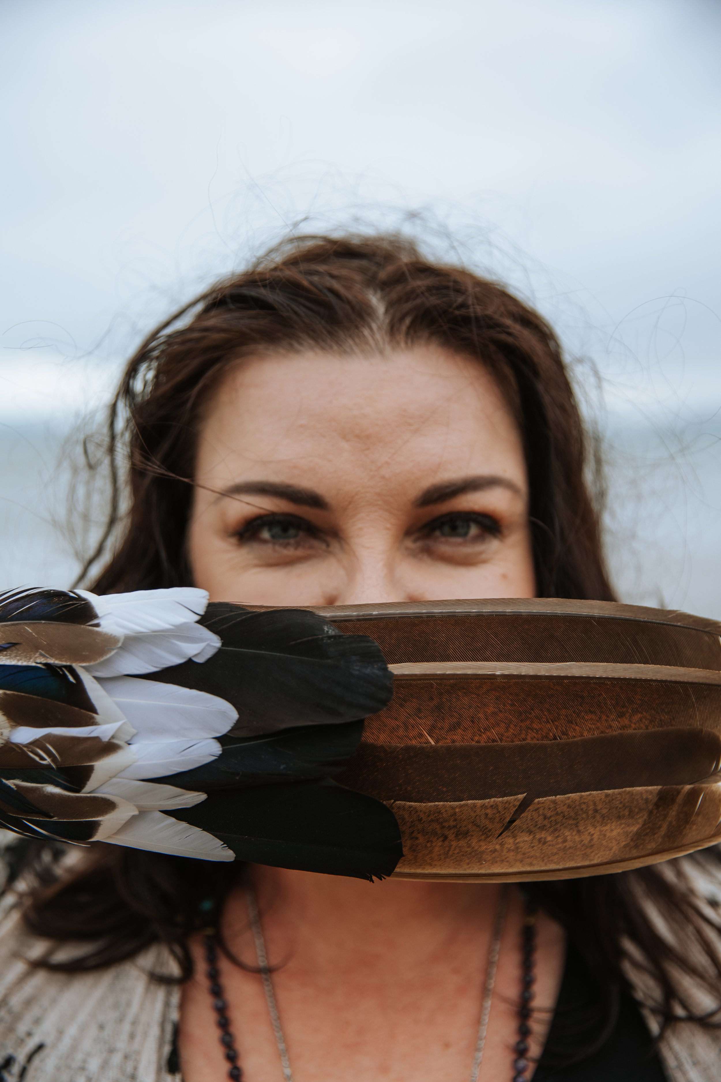 Woman holding a large bird feather close to her face outdoors, partially covering her nose and mouth, with a cloudy sky in the background.