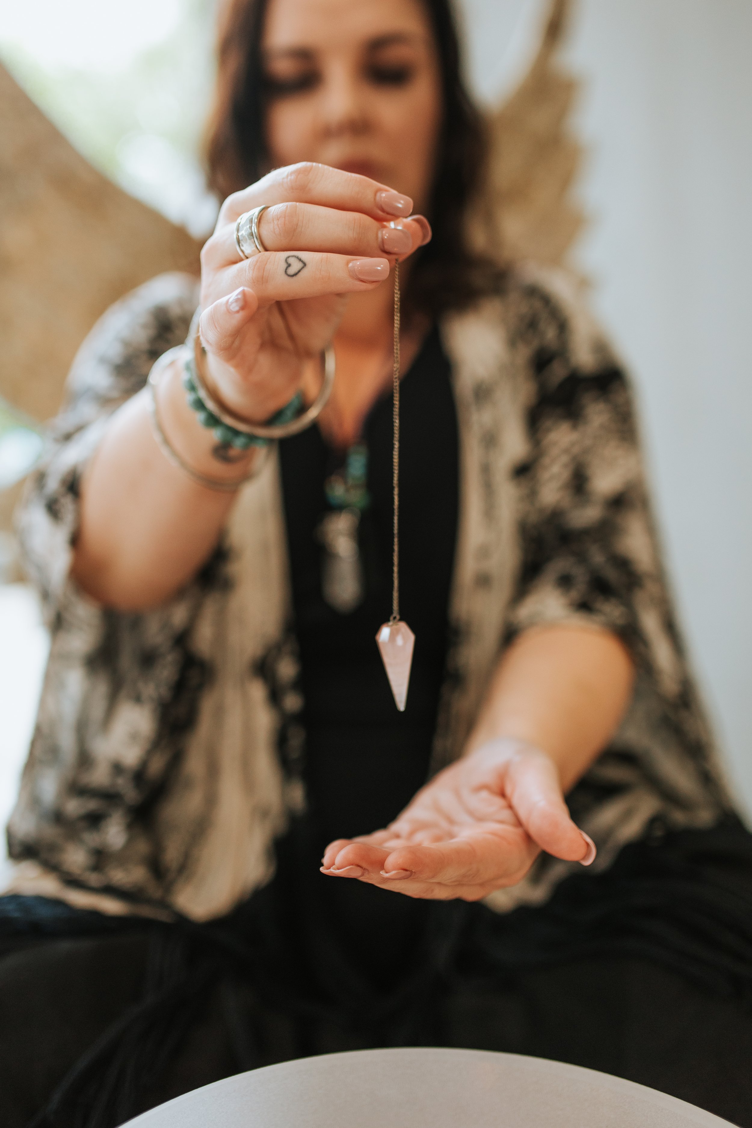 A woman holding a necklace with a pointed pendant, showing it to the camera, while seated and wearing jewelry and a patterned jacket.