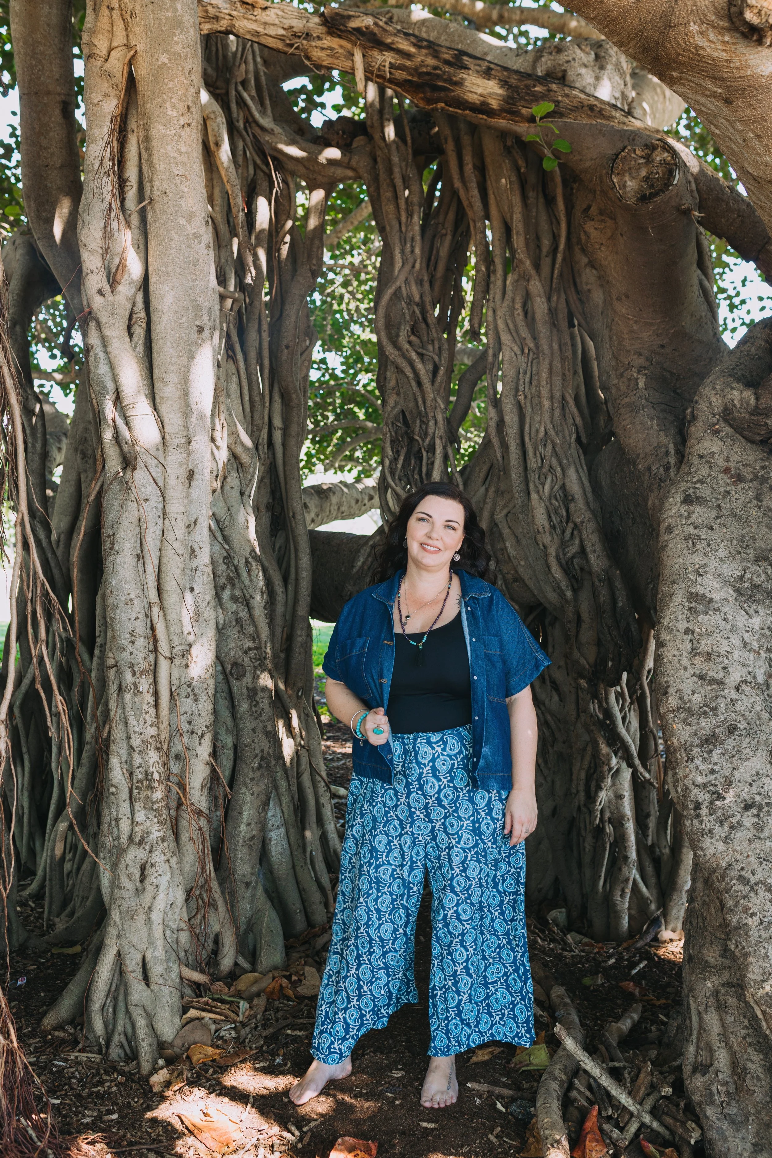A woman standing barefoot in front of a large tree with twisted roots and branches, outdoors on a sunny day.