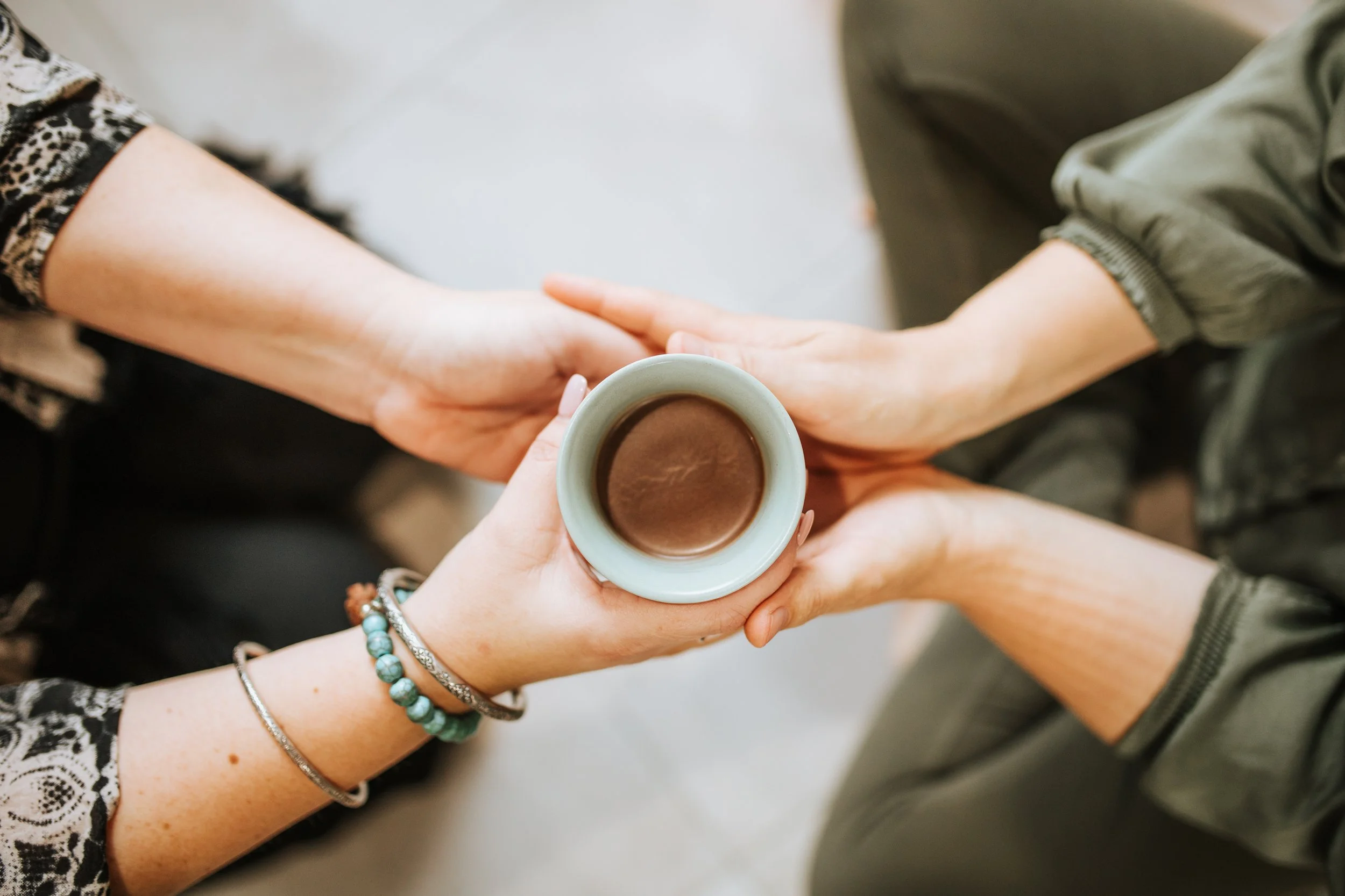 Two people exchanging a small cup of coffee or hot chocolate, holding it together with both hands, one person wearing bracelets on their wrist.