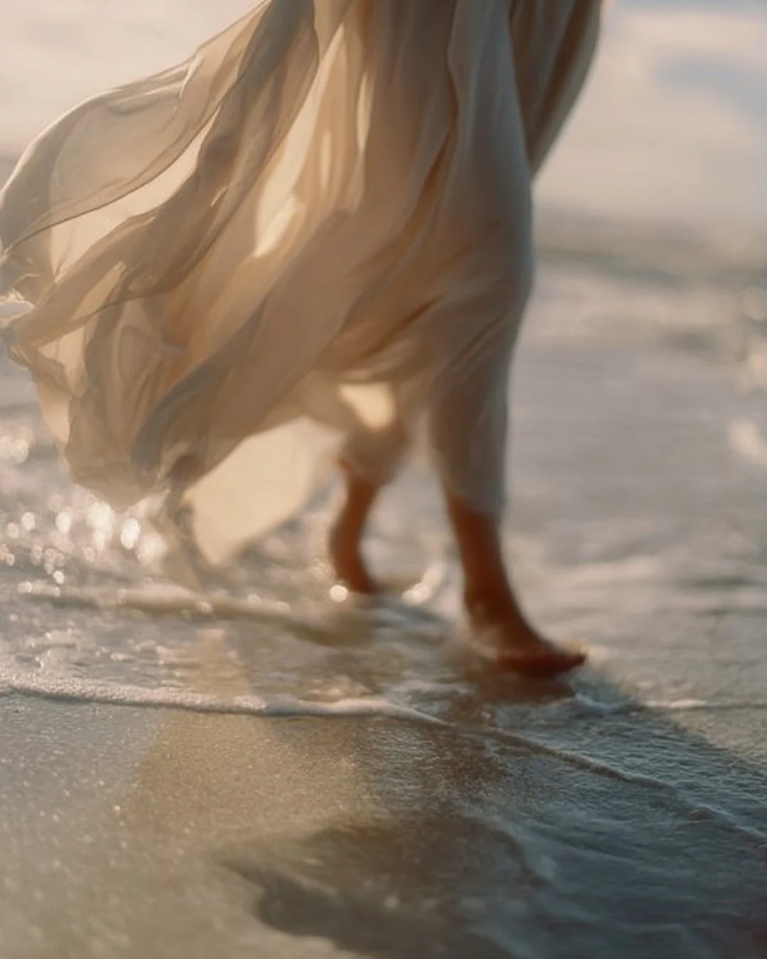 Close-up of a person walking in shallow water along the beach, with a flowing beige dress and wet sand beneath their feet.