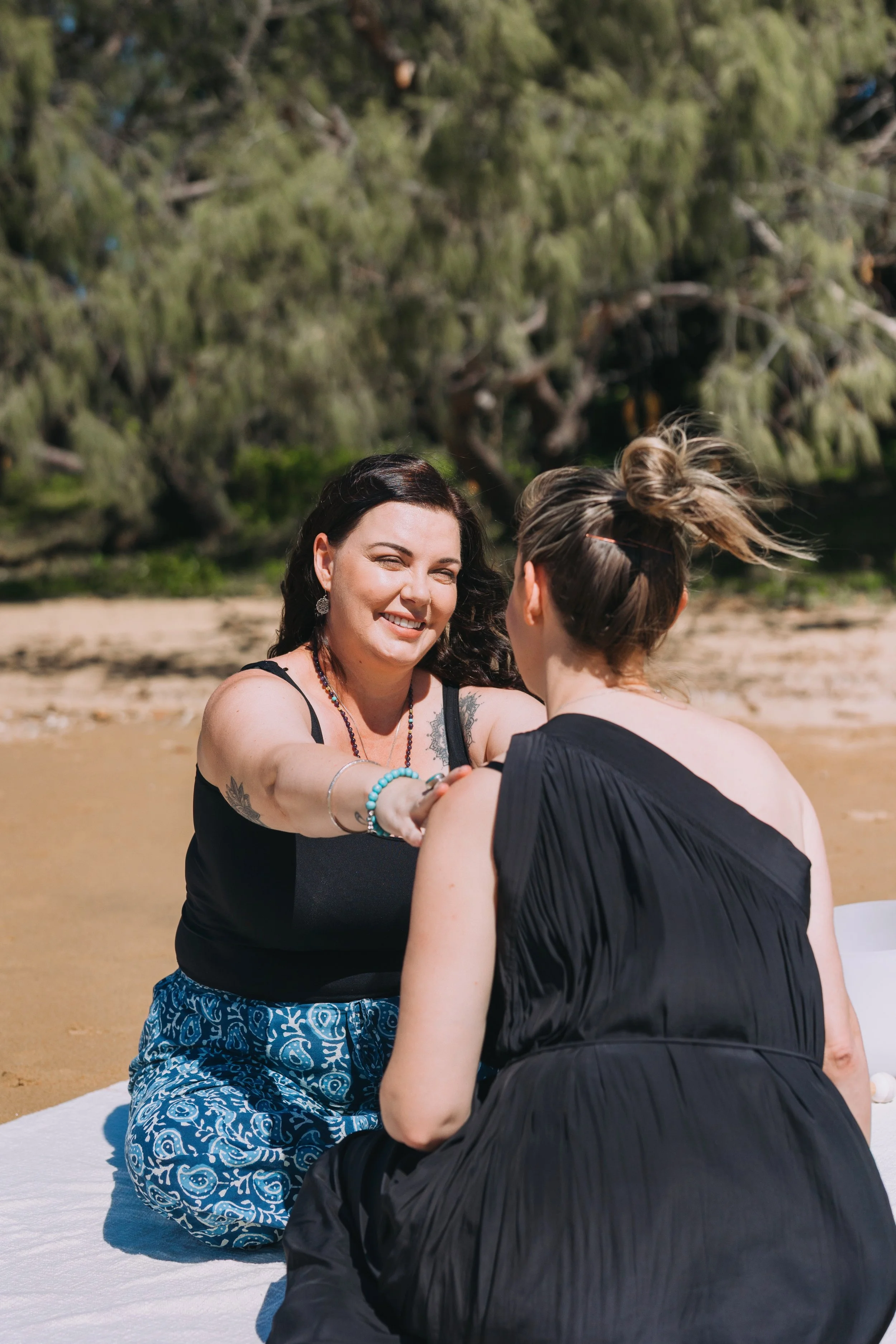 A woman smiling and extending her hand to another woman who is sitting on a beach, with trees in the background.