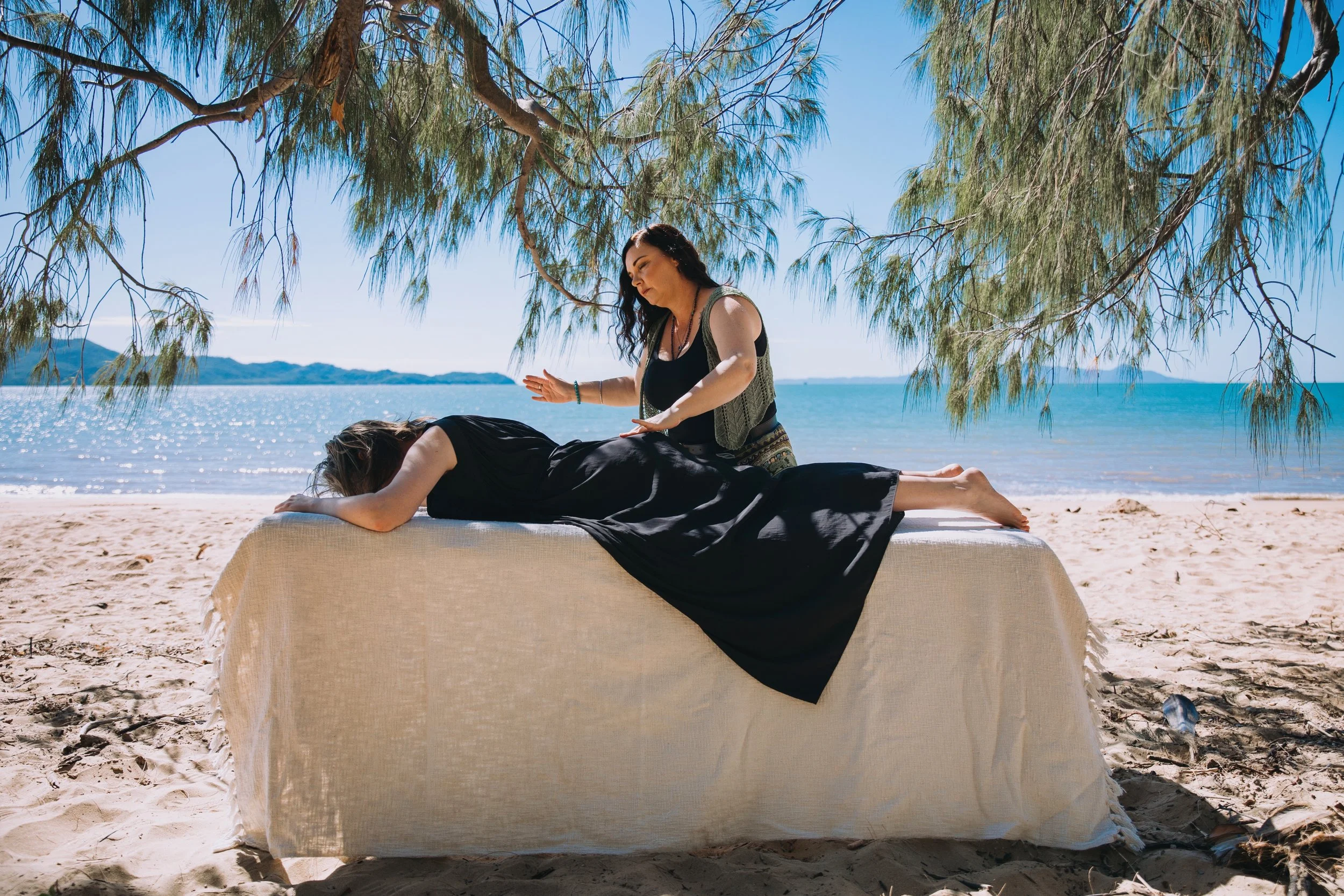A woman receives a massage on a massage table set up on a sandy beach under a tree, with the ocean and mountains in the background.