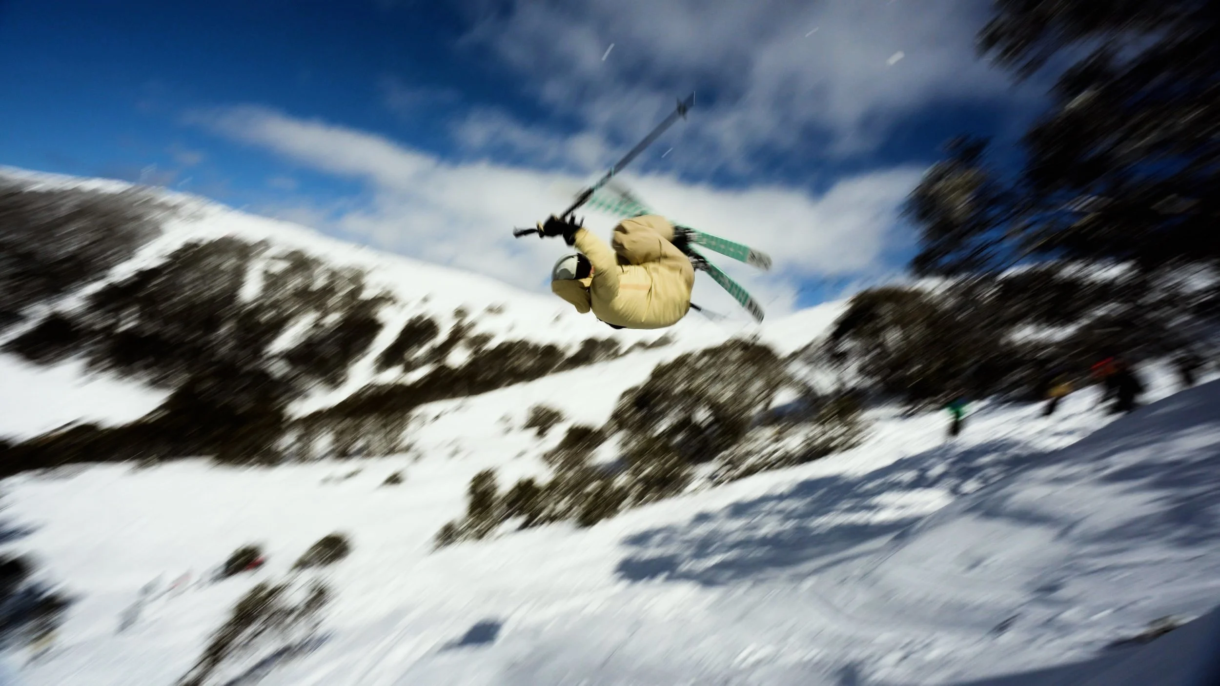 A skier in mid-air performing a jump on a snowy mountain slope with a blue sky and scattered clouds overhead.