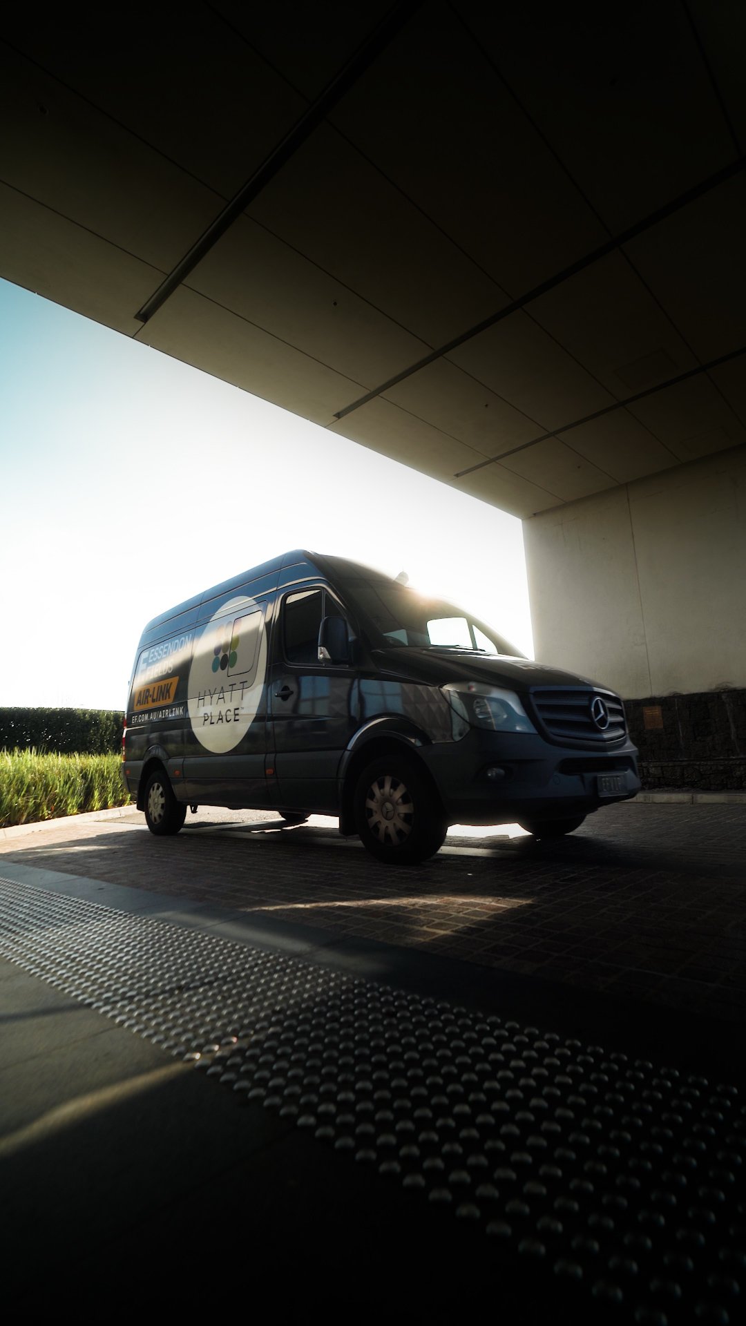 A dark-colored Mercedes-Benz van parked under a modern building structure with sunlight shining behind it, and a grassy area with trees in the background.