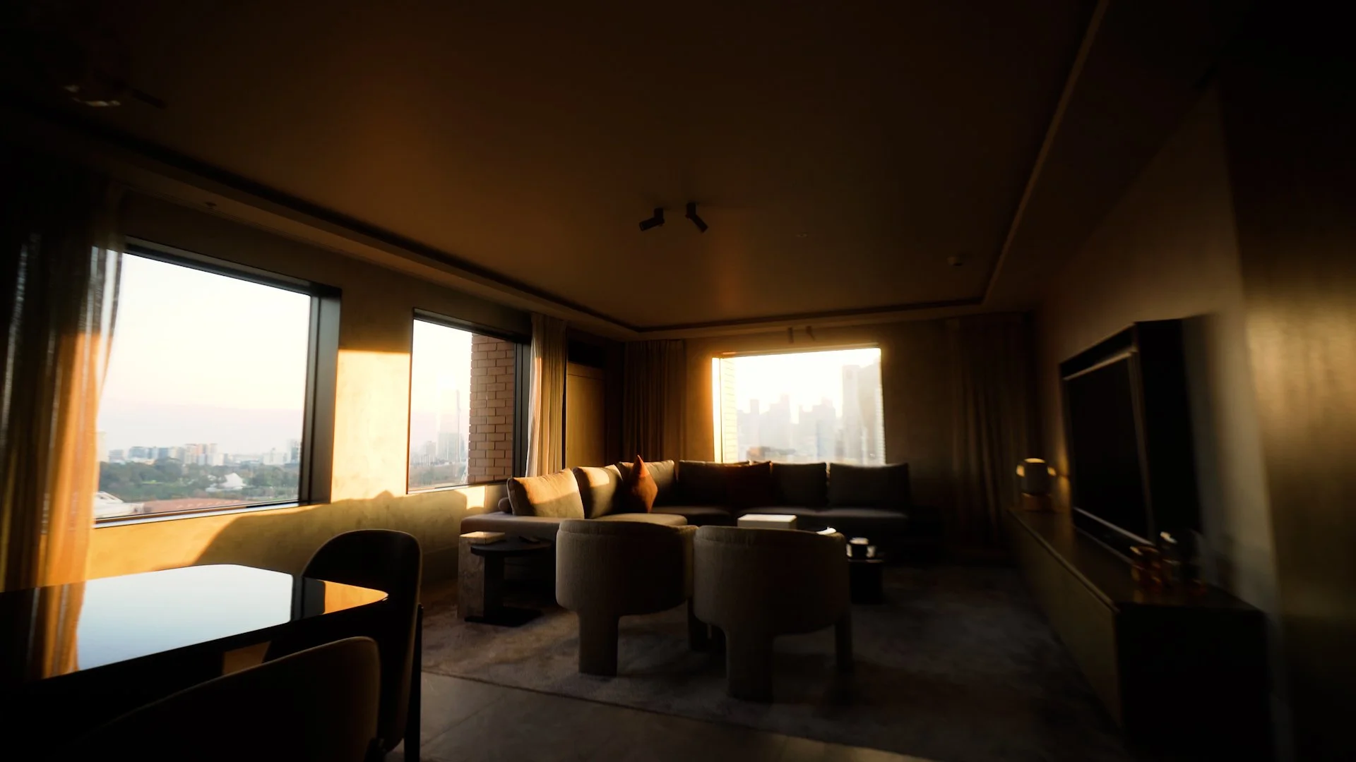 Living room with large windows showing city skyline at sunset, brown sectional sofa, armchair, television, and curtains.