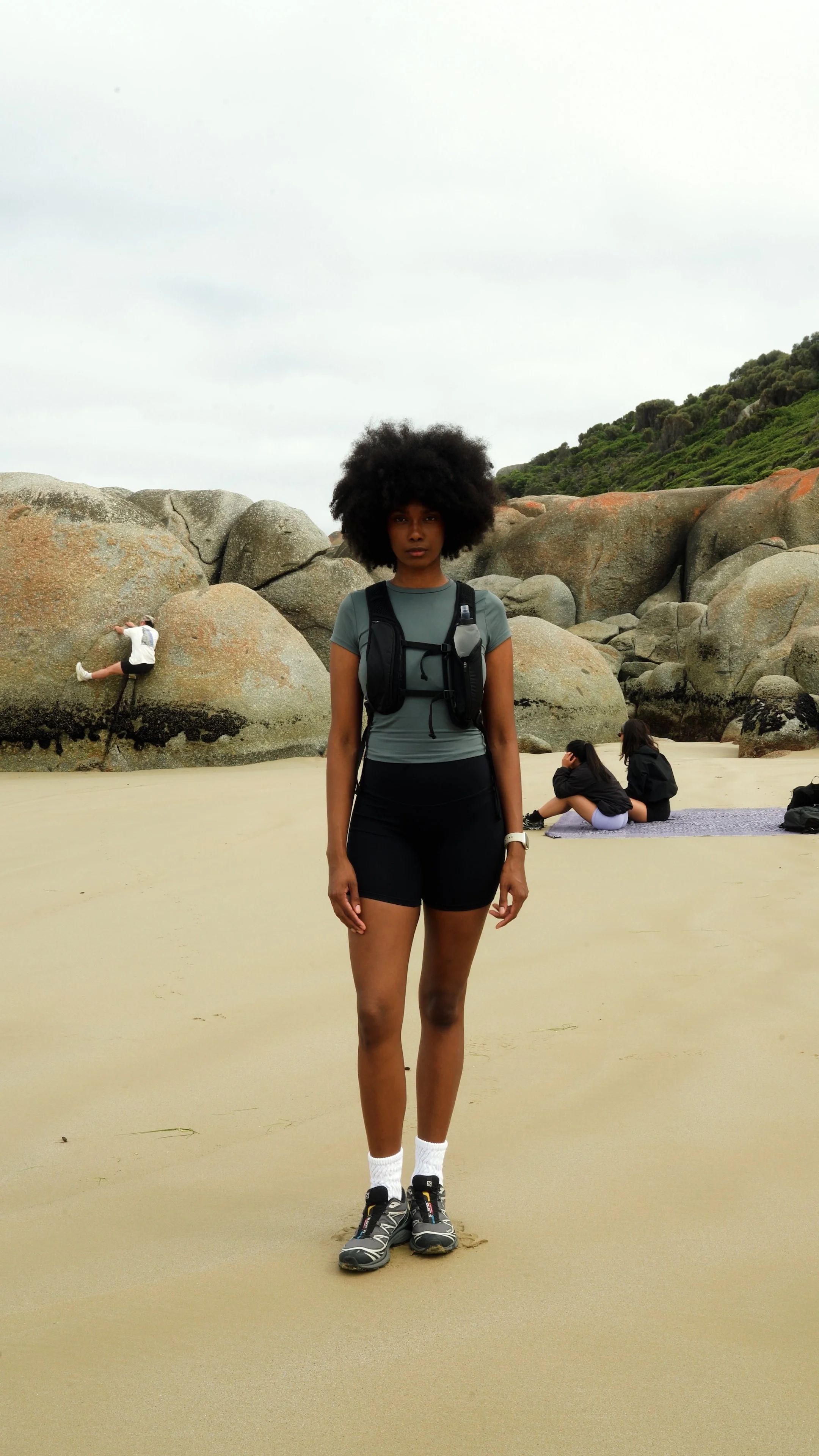 Young woman with large curly hair standing on a sandy beach, wearing athletic clothes, sneakers, and a hydration pack, with other people relaxing and climbing rocks in the background.
