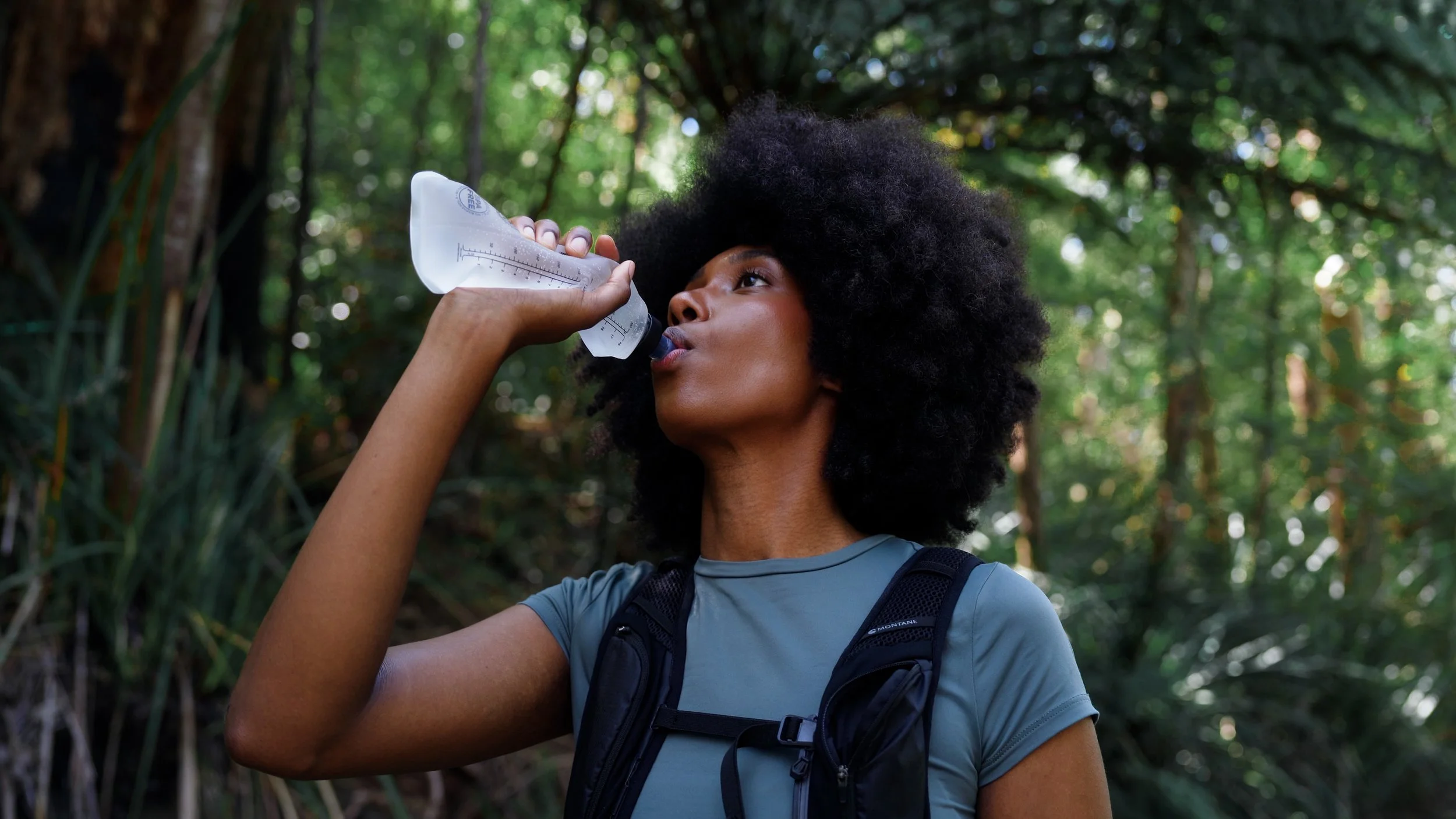 A young woman with curly black hair and athletic clothing drinks water from a hydration pack in a forest during a hike.