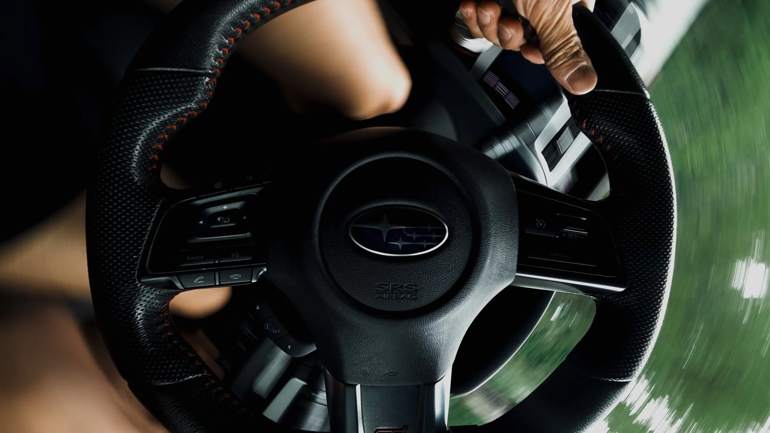 Close-up of a person's hand gripping a steering wheel inside a car moving fast, with blurred greenery outside the window.