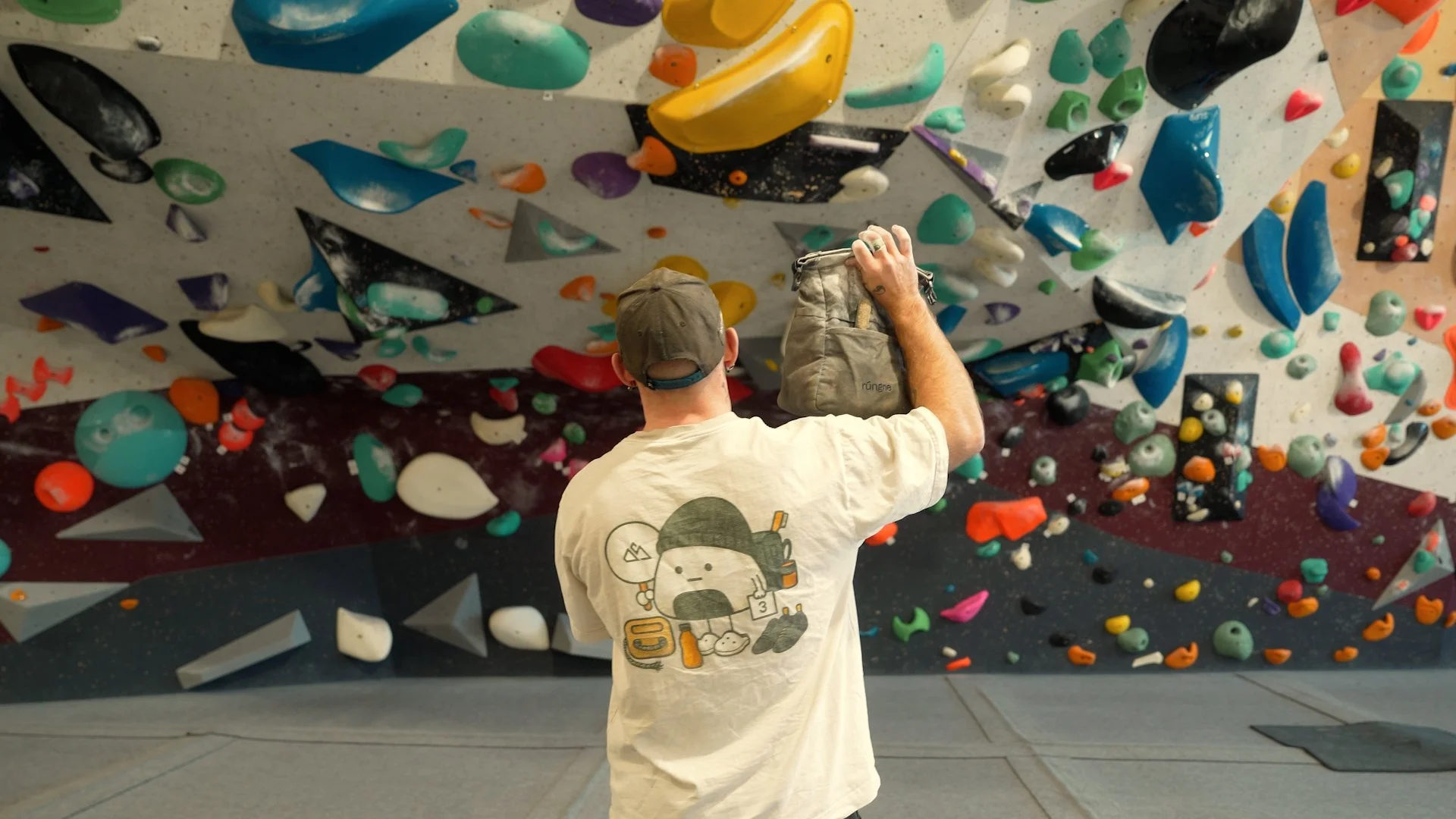 Person with a gray cap and a beige T-shirt with a cartoon character on the back is seen from behind, adjusting climbing holds on an indoor bouldering wall covered with colorful holds.