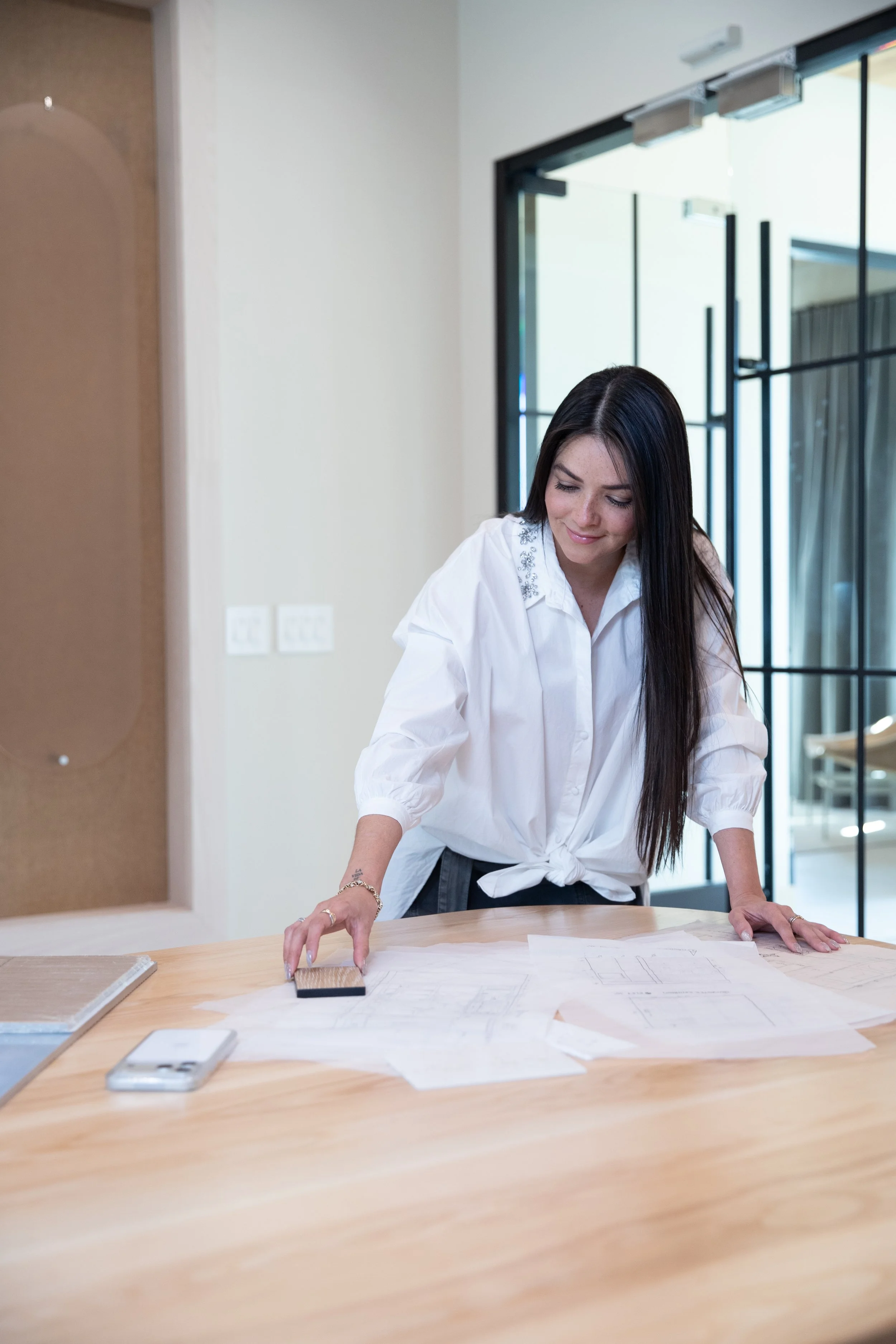 Woman leaning over a wooden table, working on architectural plans or blueprints, with a swatch in her hand, in a modern office or studio space.