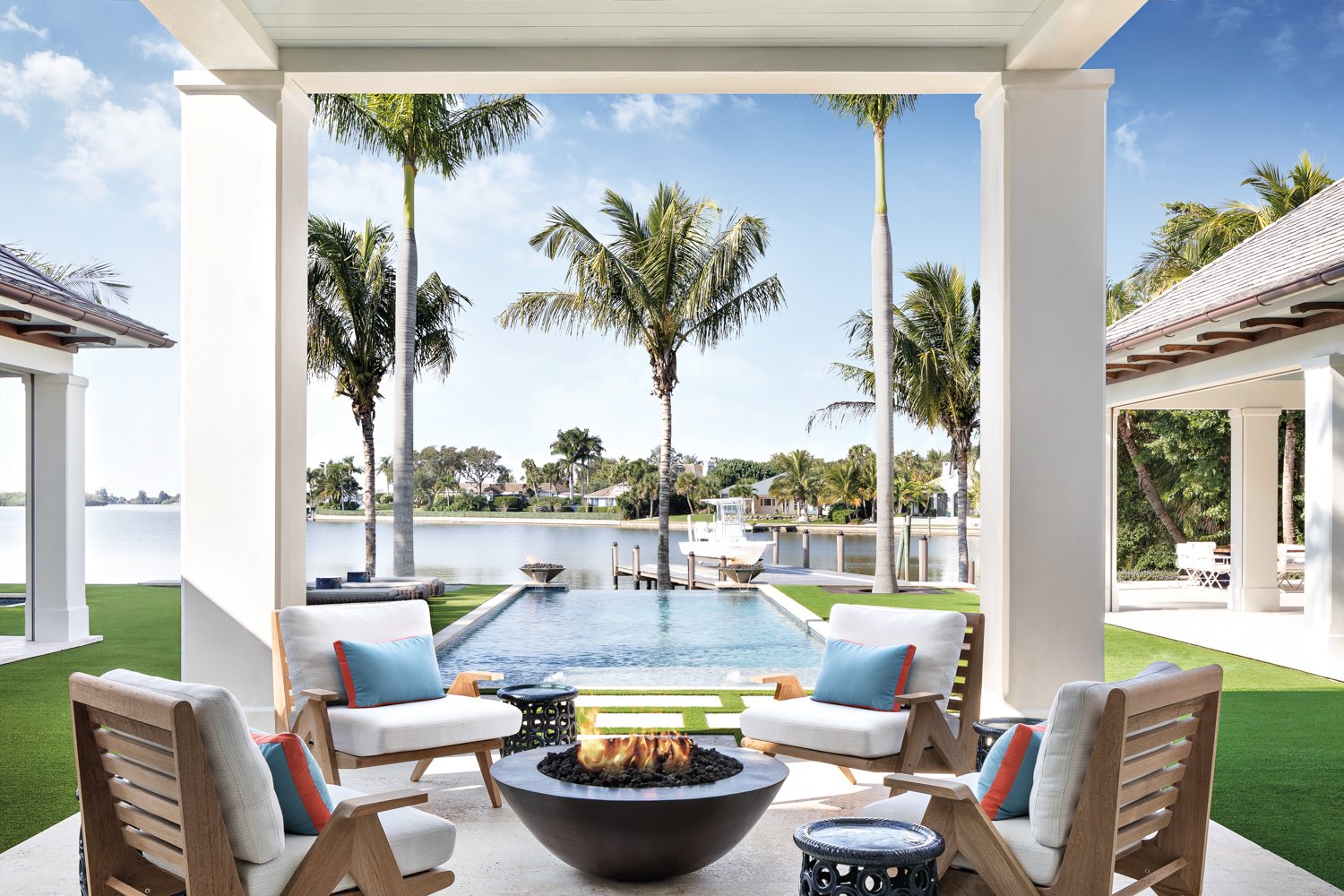 Outdoor patio with four cushioned chairs around a firepit, overlooking a waterway with palm trees and a boat dock