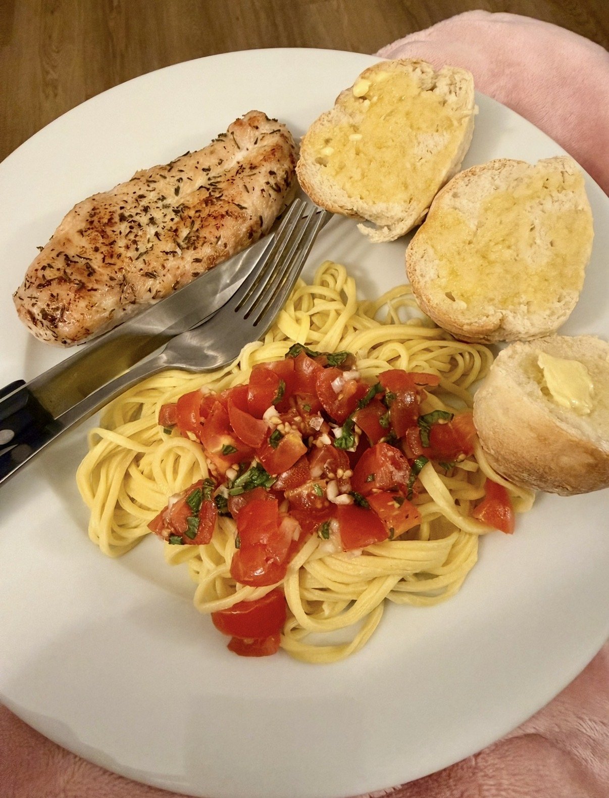 A plate of cooked spaghetti with chopped tomato and basil, grilled chicken breast with herbs, and garlic bread with butter.