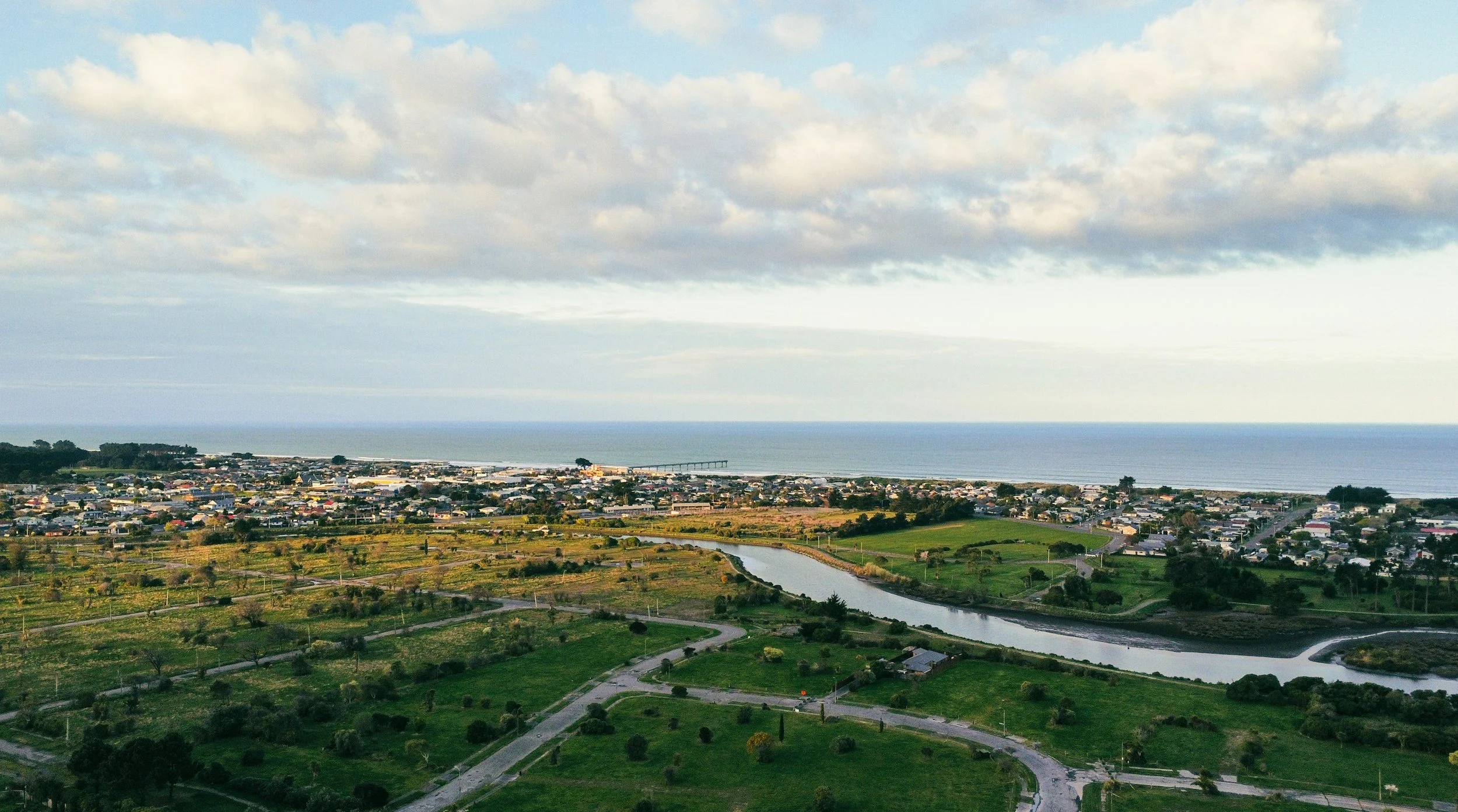 Aerial view of a coastal town with a pier extending into the ocean, a river running through green fields, and a sky with scattered clouds.