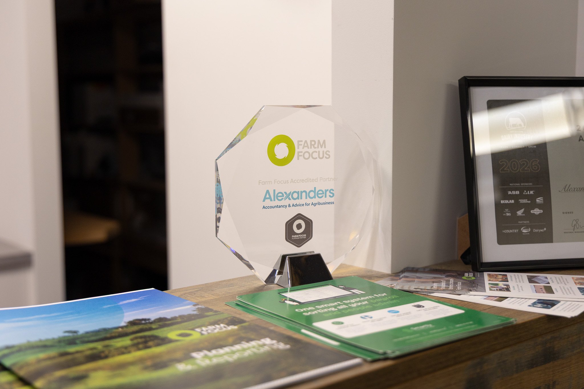 A clear glass award with a green Farm Focus logo and the name Alexanders, placed on a table among farm-related brochures and pamphlets. There is a framed certificate or award and some papers nearby.