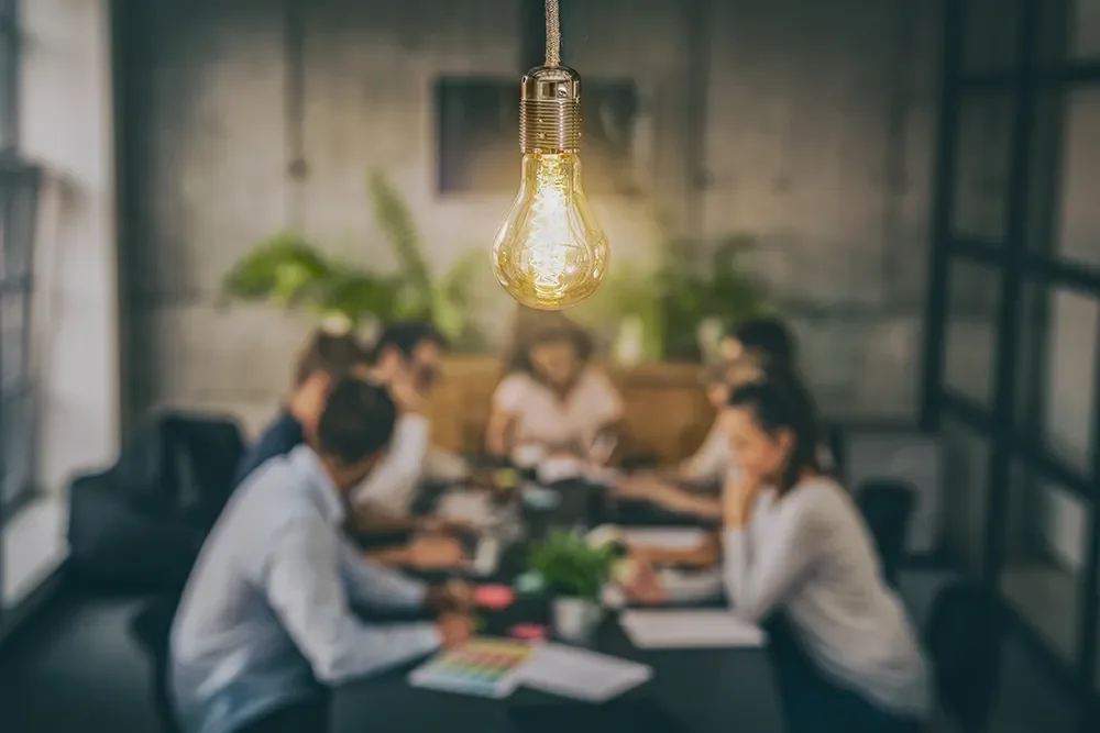 Group of people in a meeting room with a glowing light bulb hanging overhead.