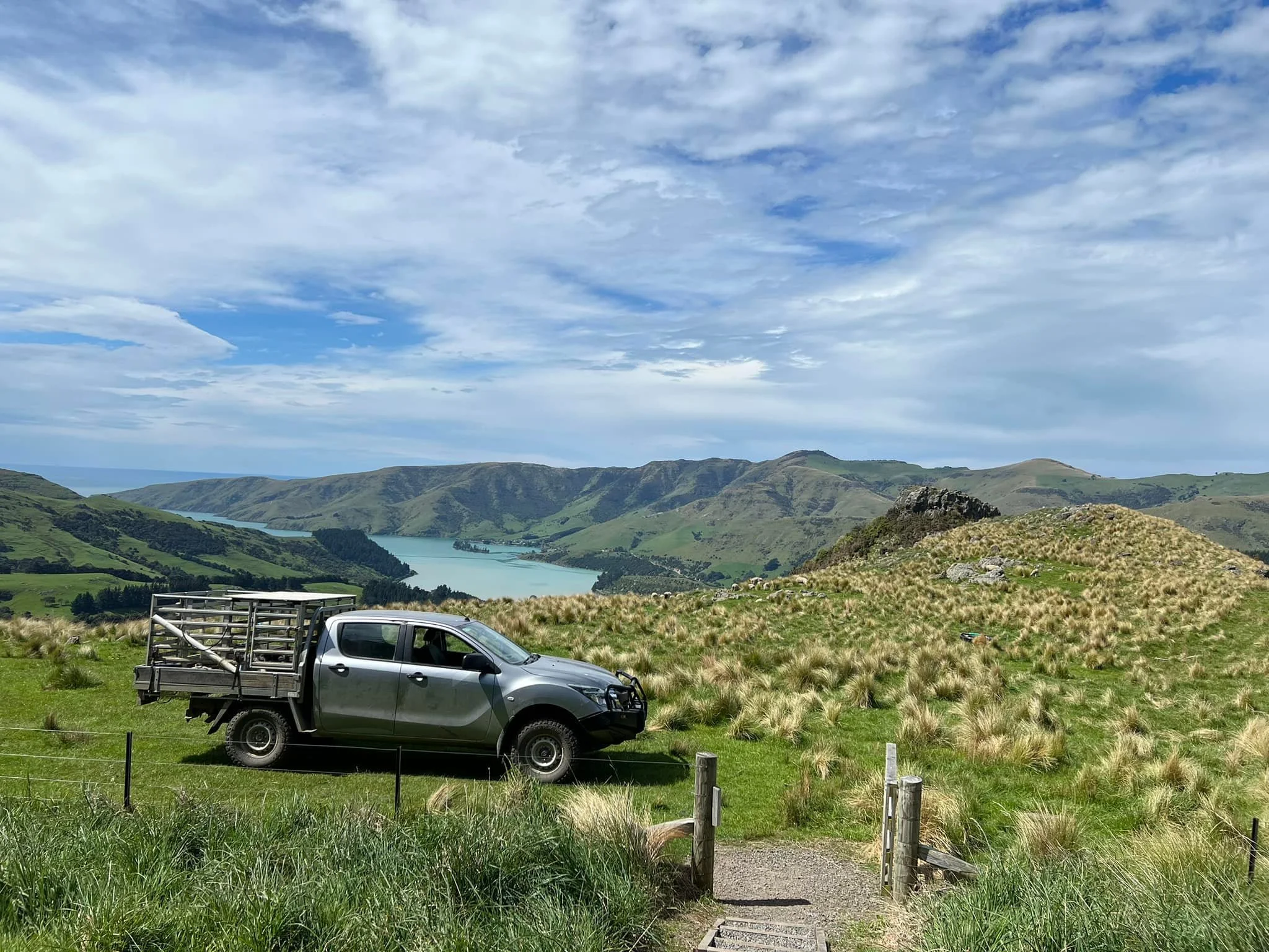 A landscape view of rolling green hills with a reservoir lake in the middle, under a partly cloudy sky. In the foreground, a gray pickup truck with a cargo rack on the back is parked on grass, near a small wooden fence and a dirt pathway.