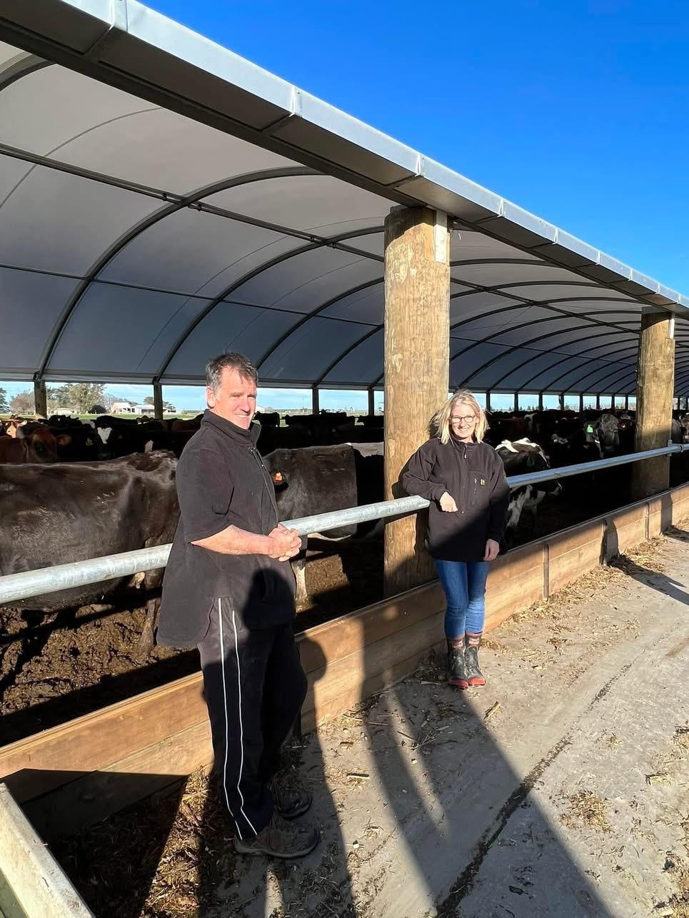 Two people, a man and a woman, standing by a metal fence at a cattle ranch with a covered shelter. The man is wearing a black jacket and black pants with stripes, and the woman is wearing a black hoodie, jeans, and boots. Behind them are cattle inside the shelter, and the sky is clear and blue.