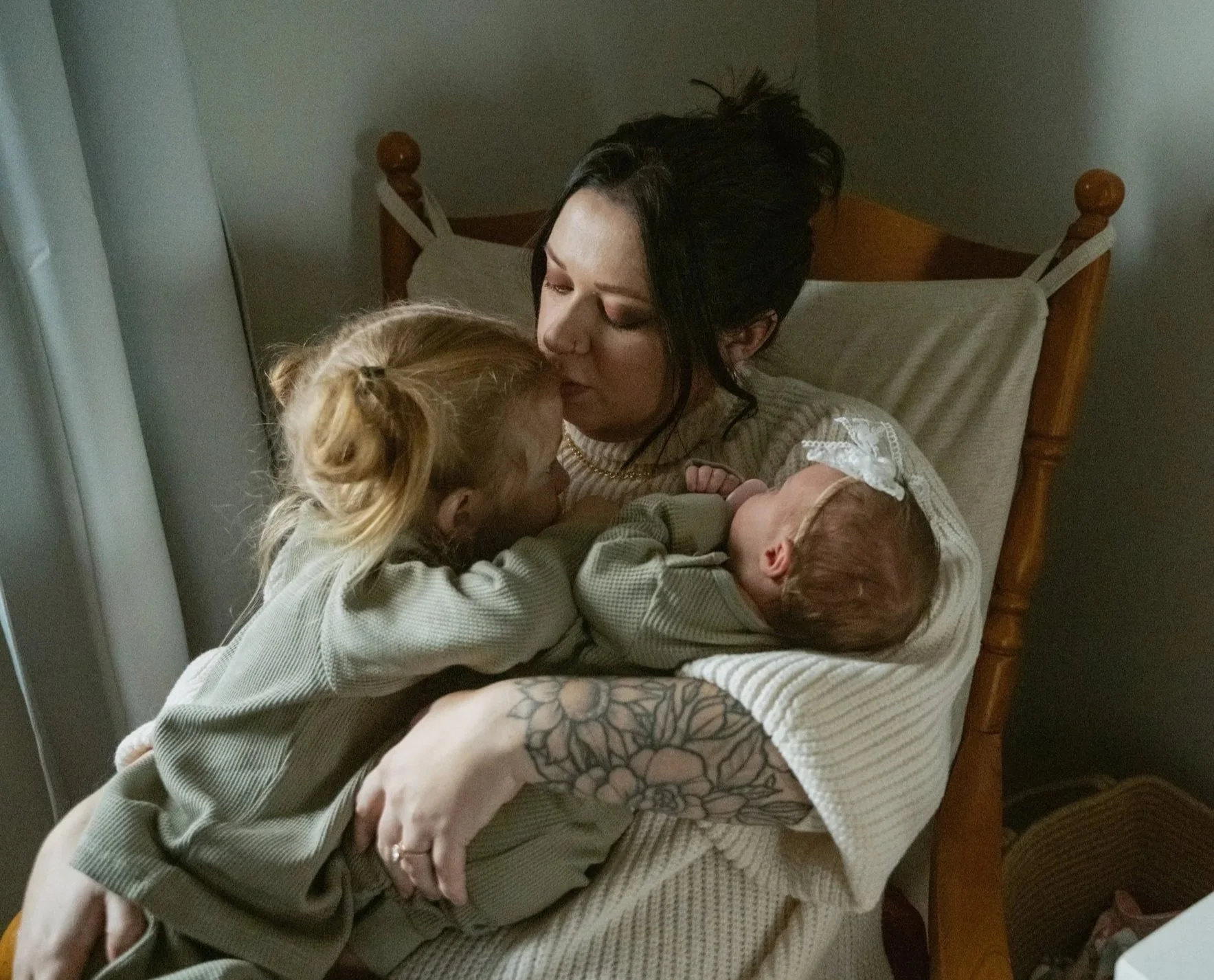 A woman with black hair and tattoos on her arm appears to be holding and kissing a young child with blond hair, who is resting on her lap, with another baby with light hair nestled in her arms, all sitting on a bed with beige bedding and a wooden headboard.