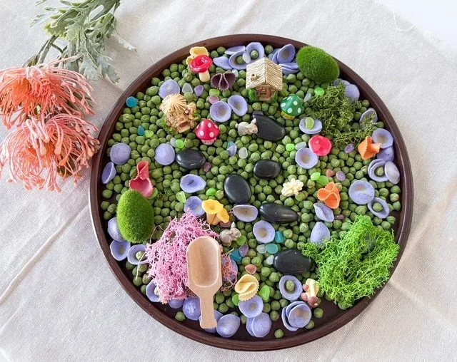 A circular tray filled with small green and purple plant beads, decorative stones, and miniature garden items like a tiny house, mushroom, and moss, surrounded by real flowers and foliage.