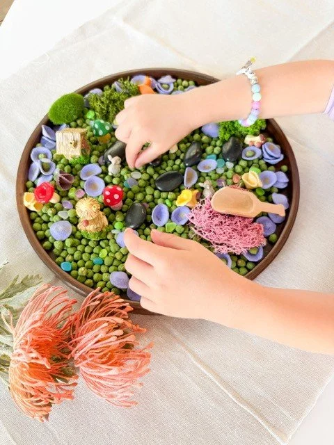Child's hands playing with colorful sensory stones and toys in a round container, with a pink flower and artificial plants nearby.