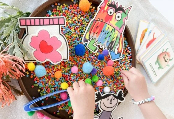 Child's hands playing with colorful sprinkles and pom-poms in a brown tray, surrounded by cartoon character images and cards.