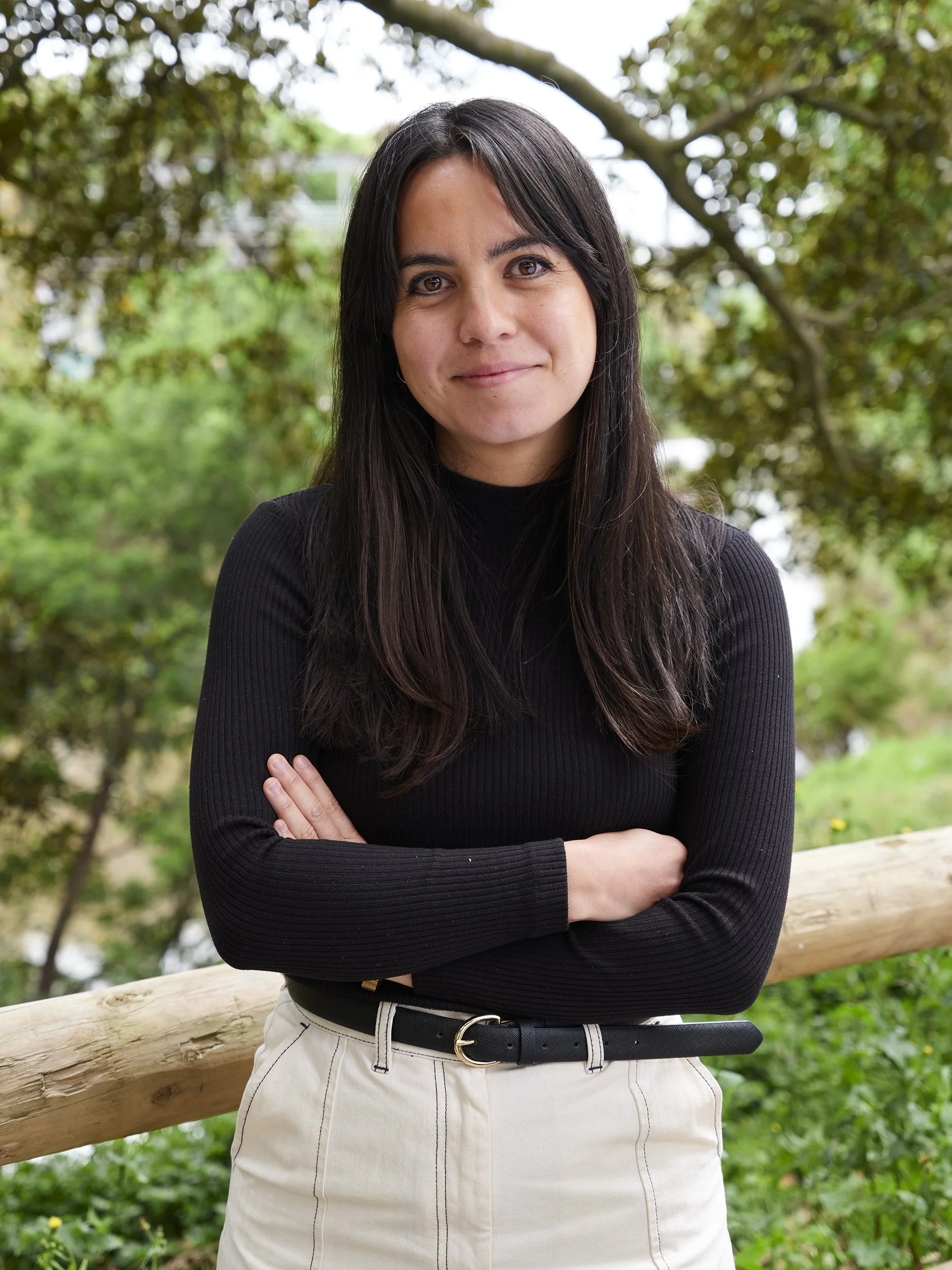 A woman with dark hair and a black long-sleeve shirt standing outdoors near a wooden fence, with green trees and foliage in the background.
