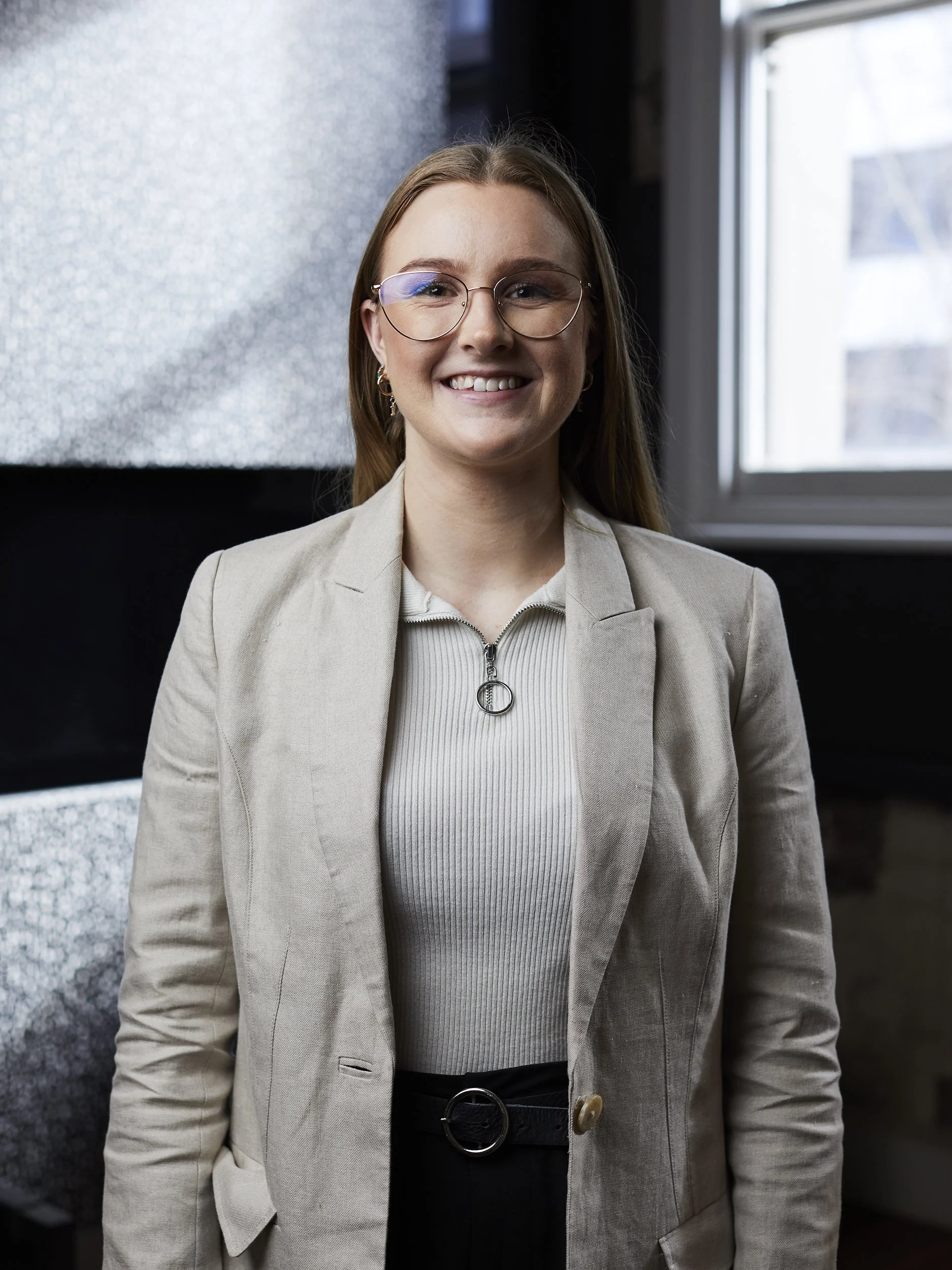 A woman smiling, wearing glasses, a beige blazer, and a light-colored shirt, standing indoors near a window.