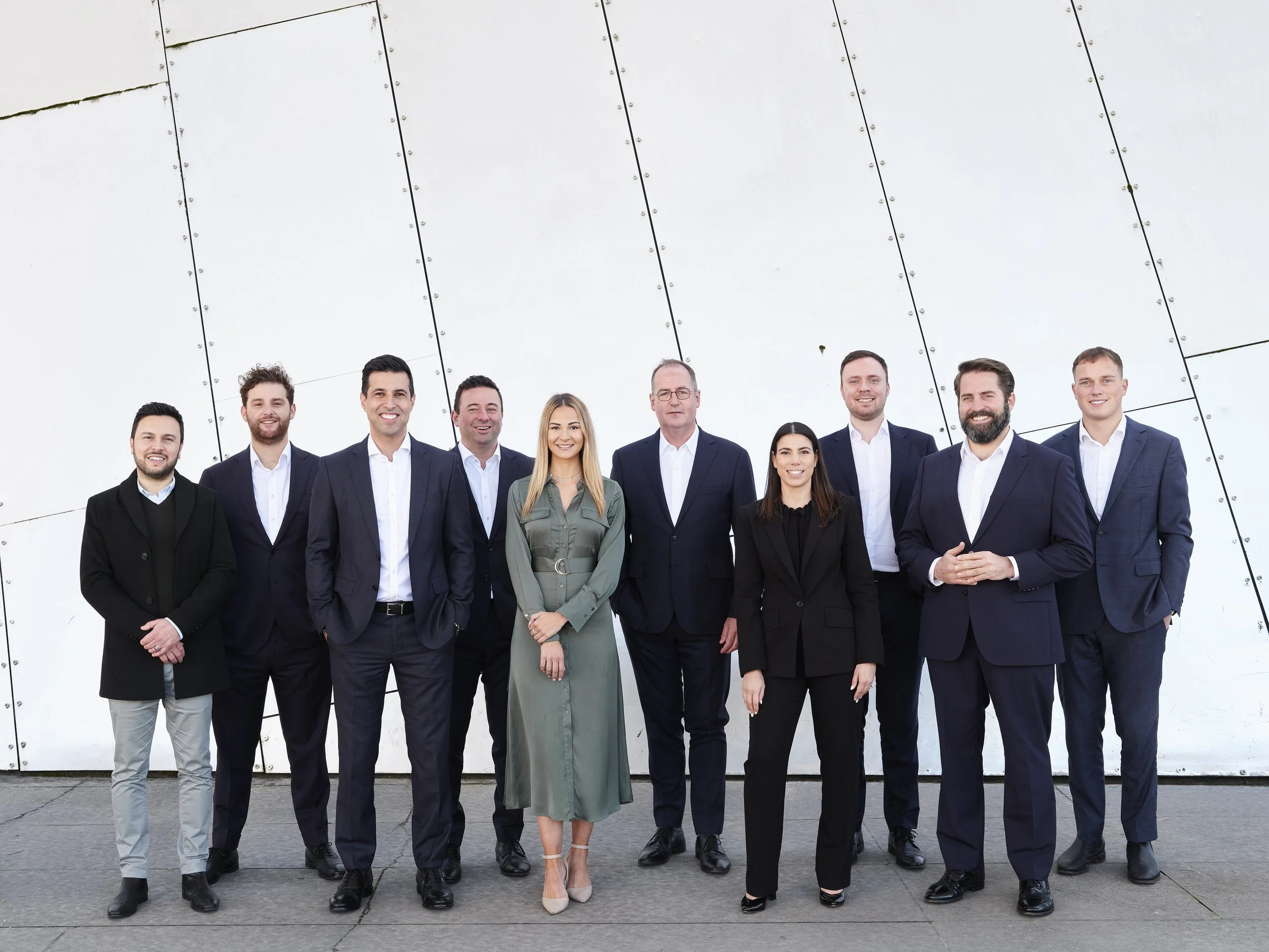 Group of ten business professionals, five men and two women, standing outdoors on a gray concrete surface in front of a geometric white wall, dressed in formal business attire, smiling at the camera.