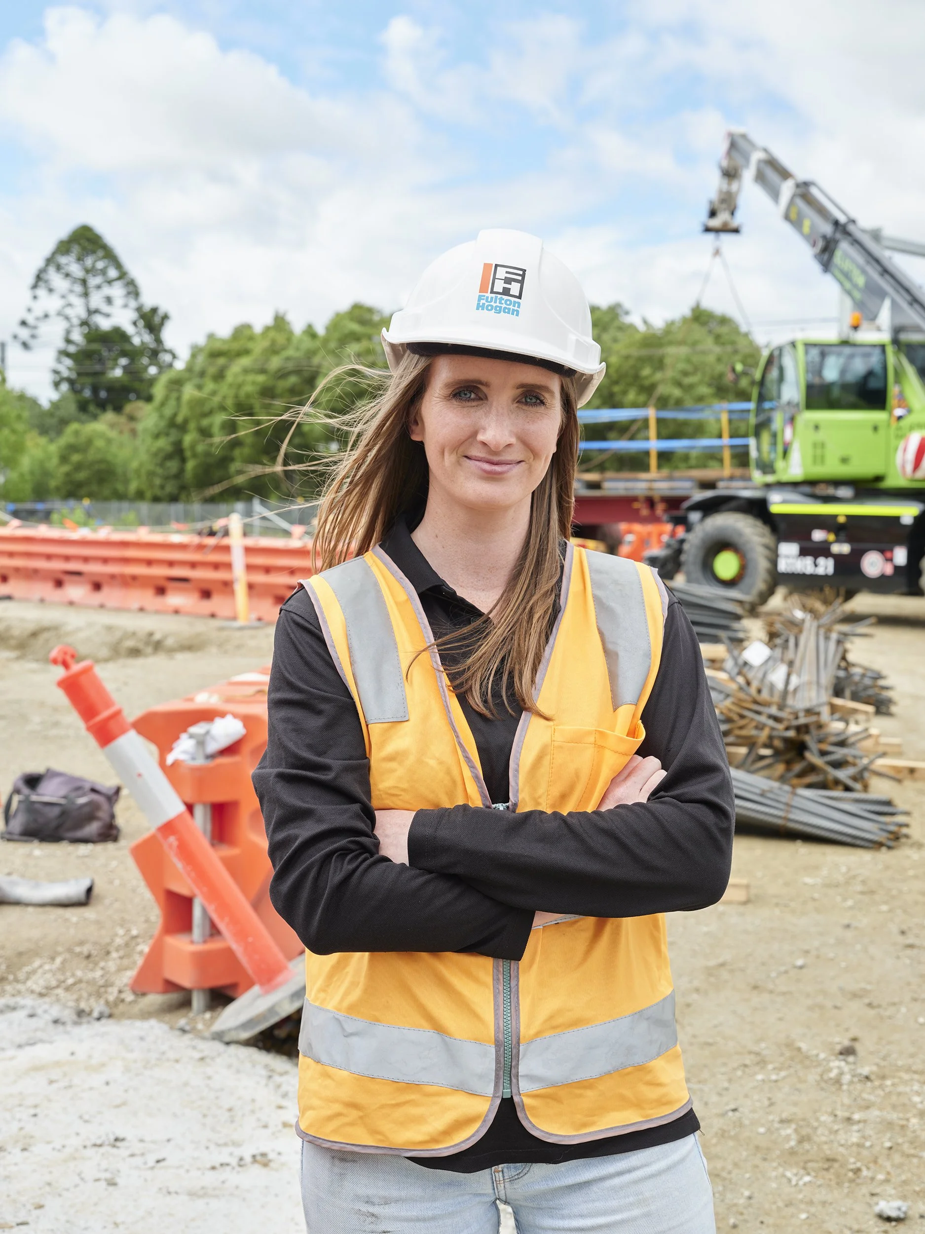 A woman in a yellow safety vest and white hard hat standing at a construction site with her arms crossed, backdropped by construction equipment and orange barriers.
