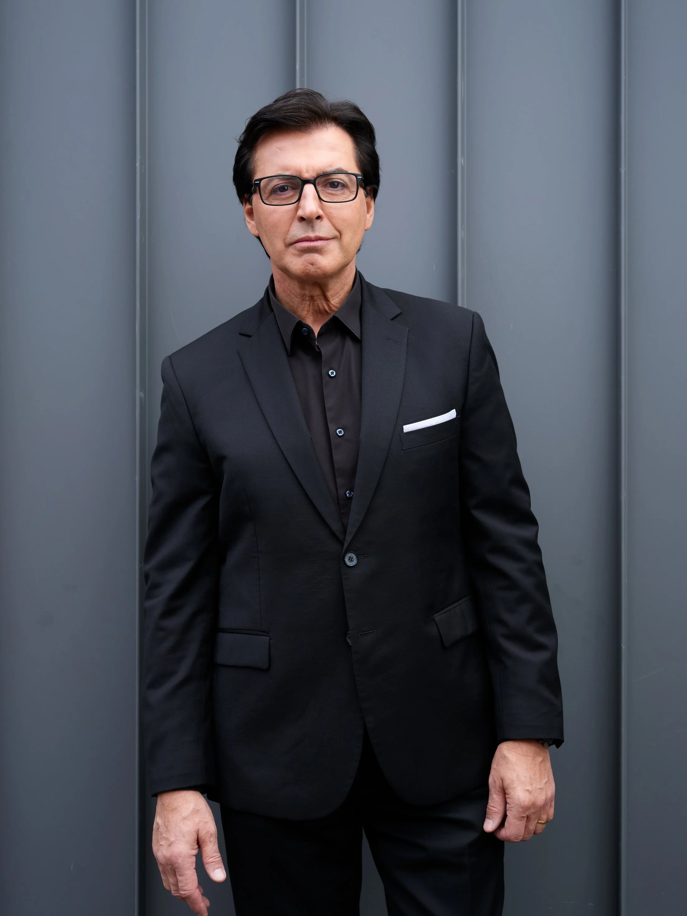 A middle-aged man with dark hair, wearing glasses, dressed in a black suit and black shirt, standing in front of a gray wall.