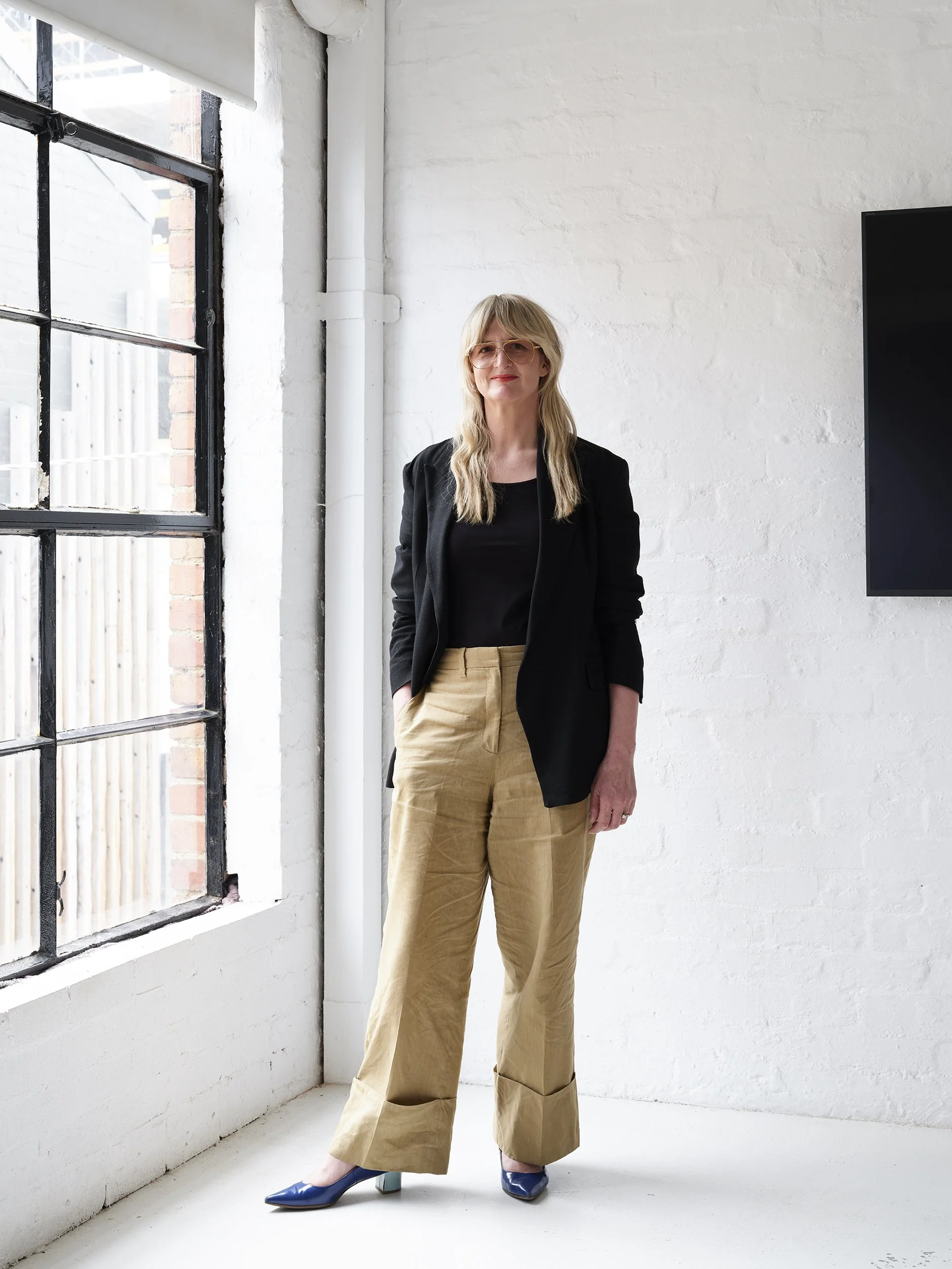 A woman with long blonde hair wearing sunglasses, a black blazer, black top, beige wide-leg pants, and blue heels standing in a bright, minimalist room with white brick walls and large window.