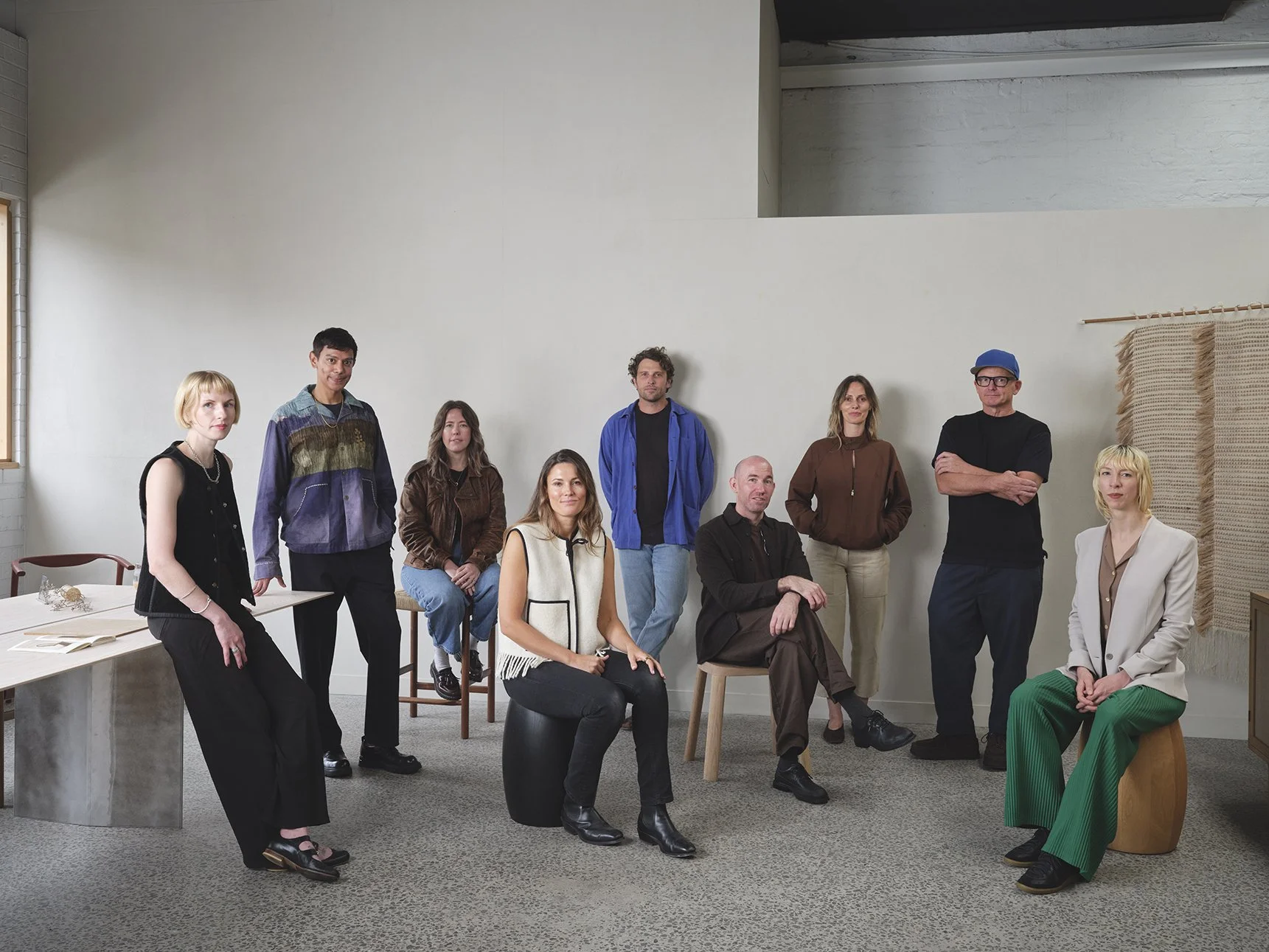 Group of nine diverse people in a modern room, some sitting on chairs or stools, others standing, posing for a photo.