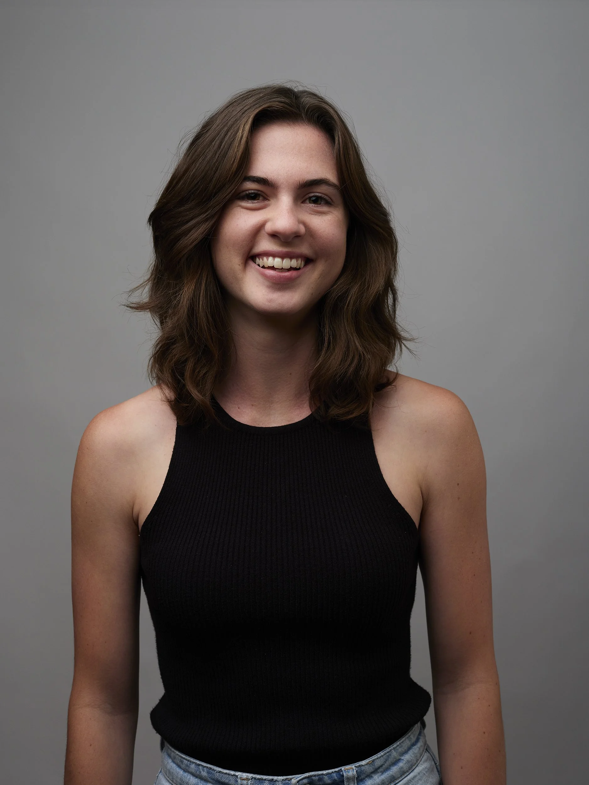 A young woman with shoulder-length brown hair, wearing a sleeveless black top and denim jeans, smiling against a plain gray background.