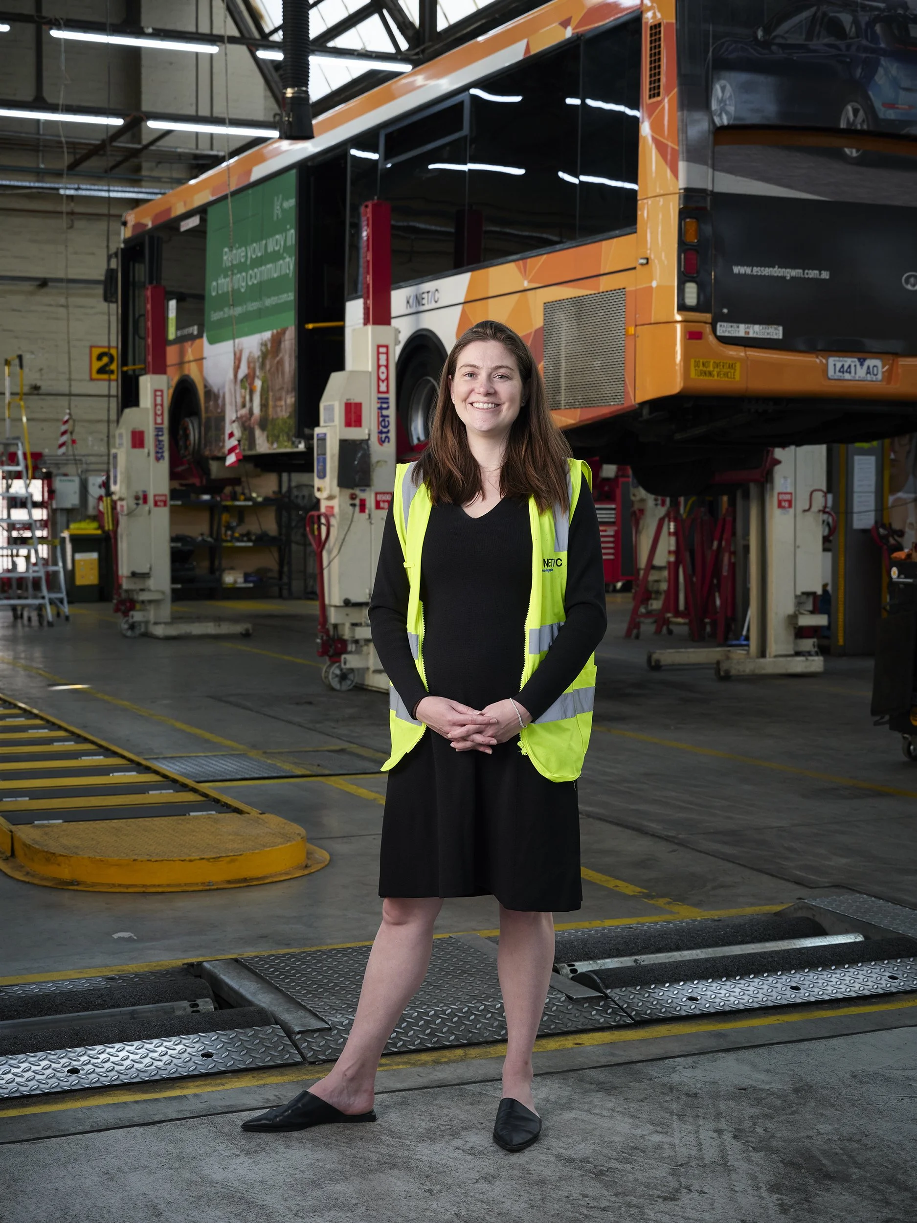 A woman wearing a black dress and a yellow safety vest standing inside an automotive workshop, with a bus raised on a hydraulic lift behind her.