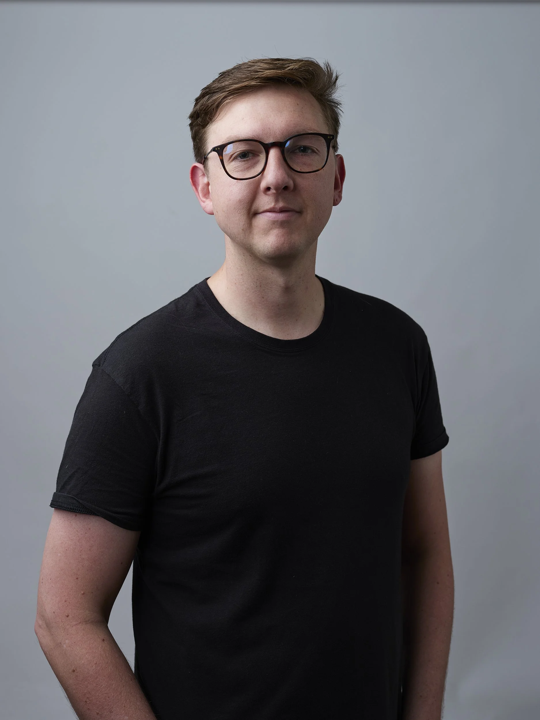 A young man with light skin and brown hair wearing glasses and a black t-shirt, standing against a plain grey background.