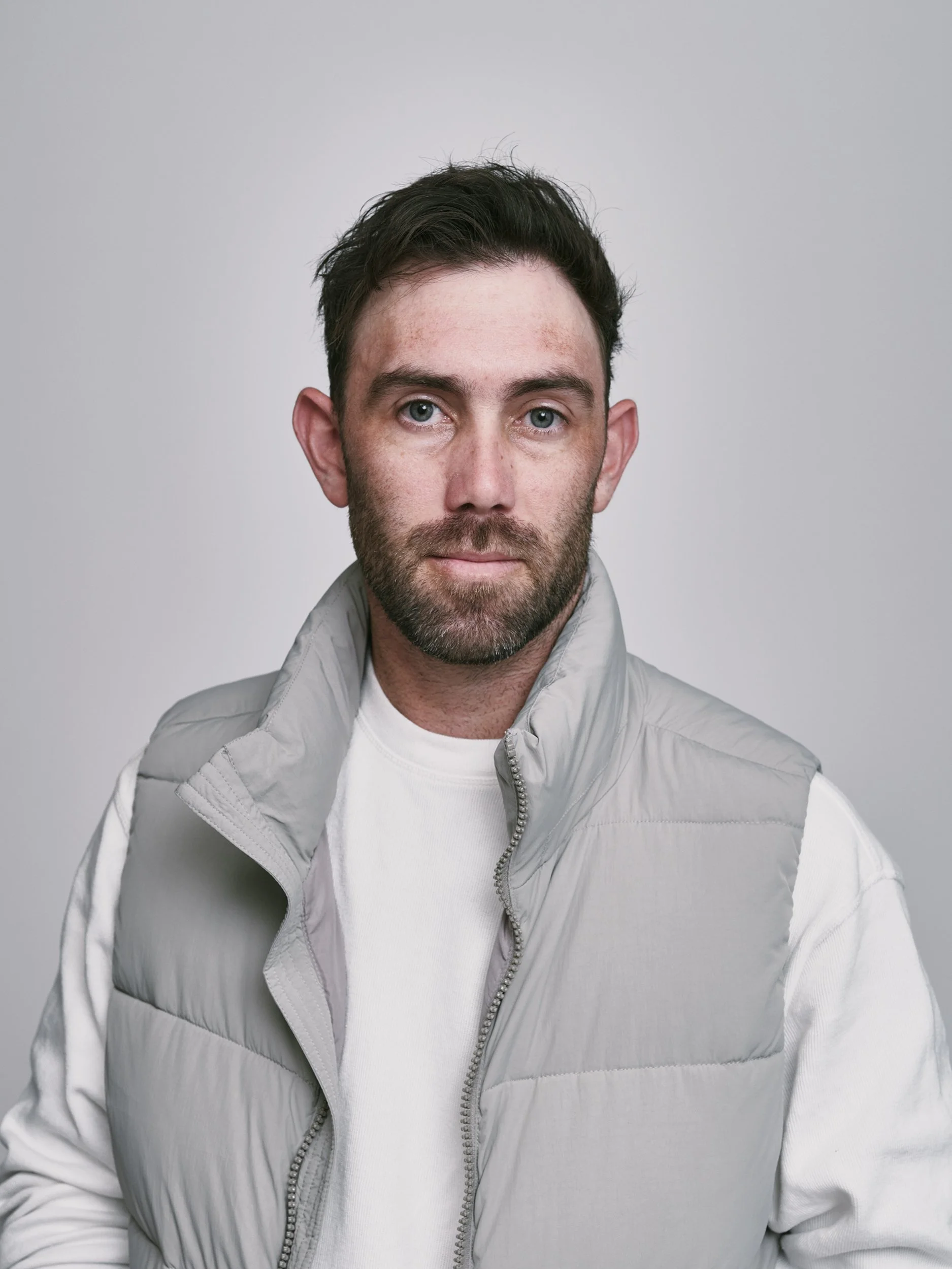 A man with dark hair and blue eyes, wearing a white shirt and a light gray padded vest, posing against a plain gray background.