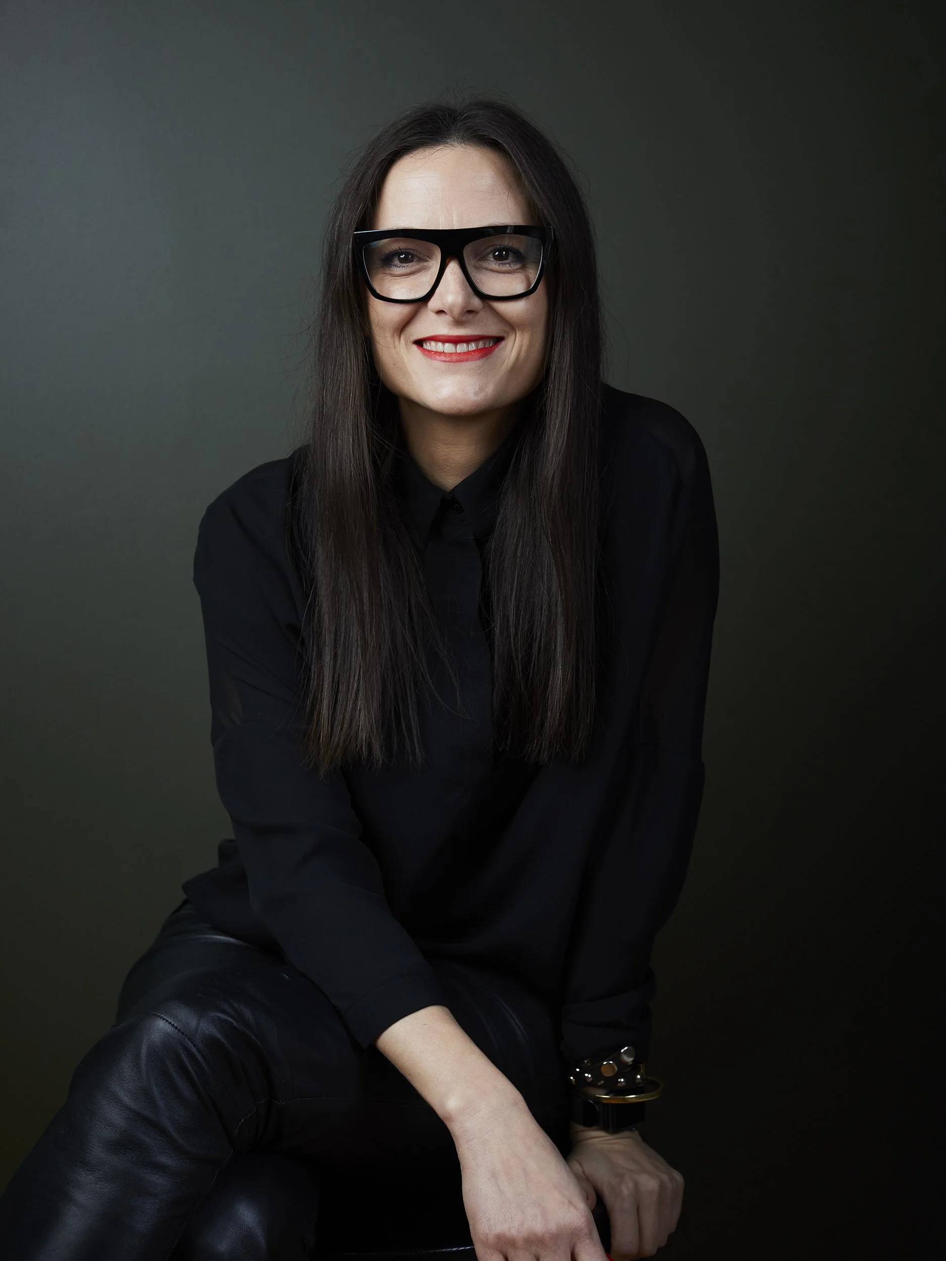 A woman with long dark hair, wearing glasses, a black shirt, and leather pants, smiling against a dark green background.