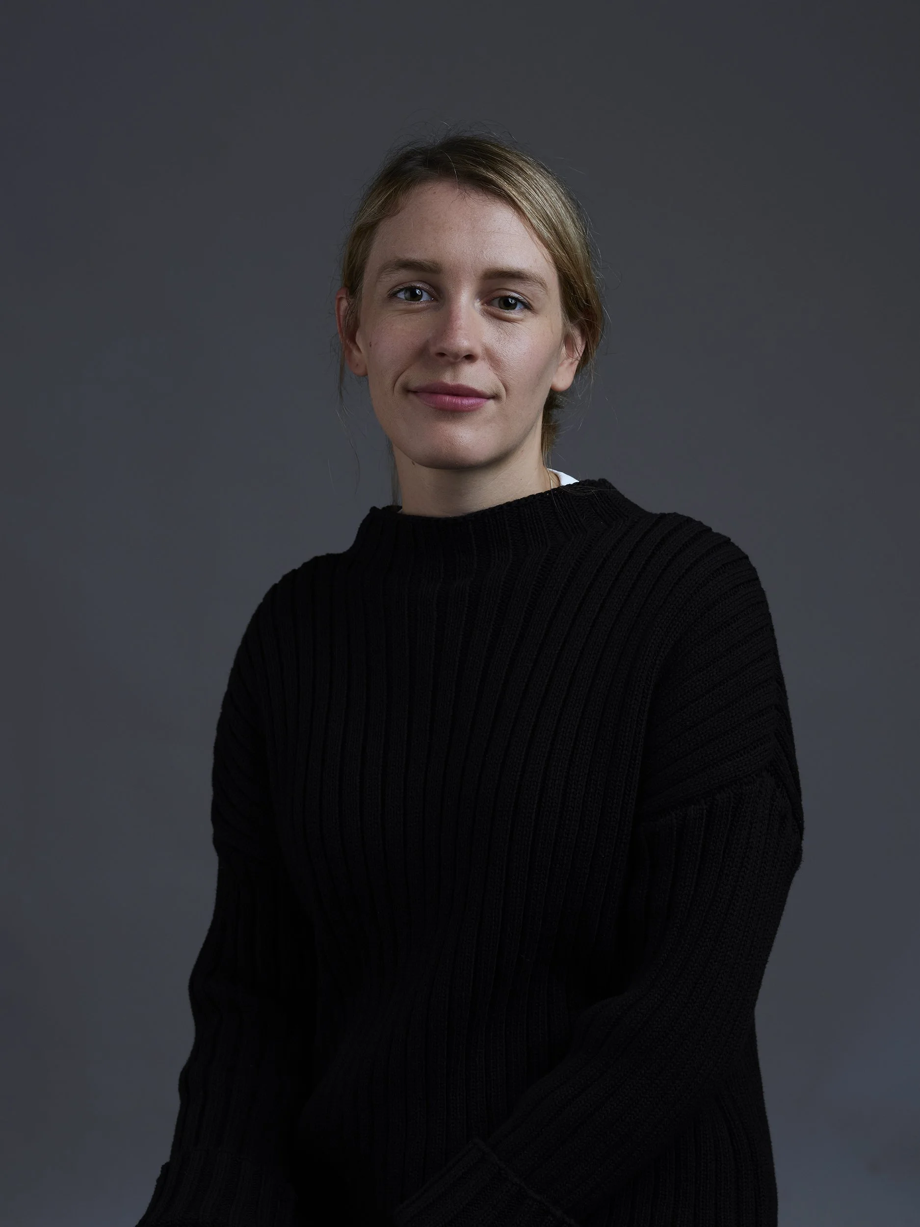 A young woman with blonde hair and fair skin, wearing a black ribbed sweater, standing against a gray background with a slight smile.