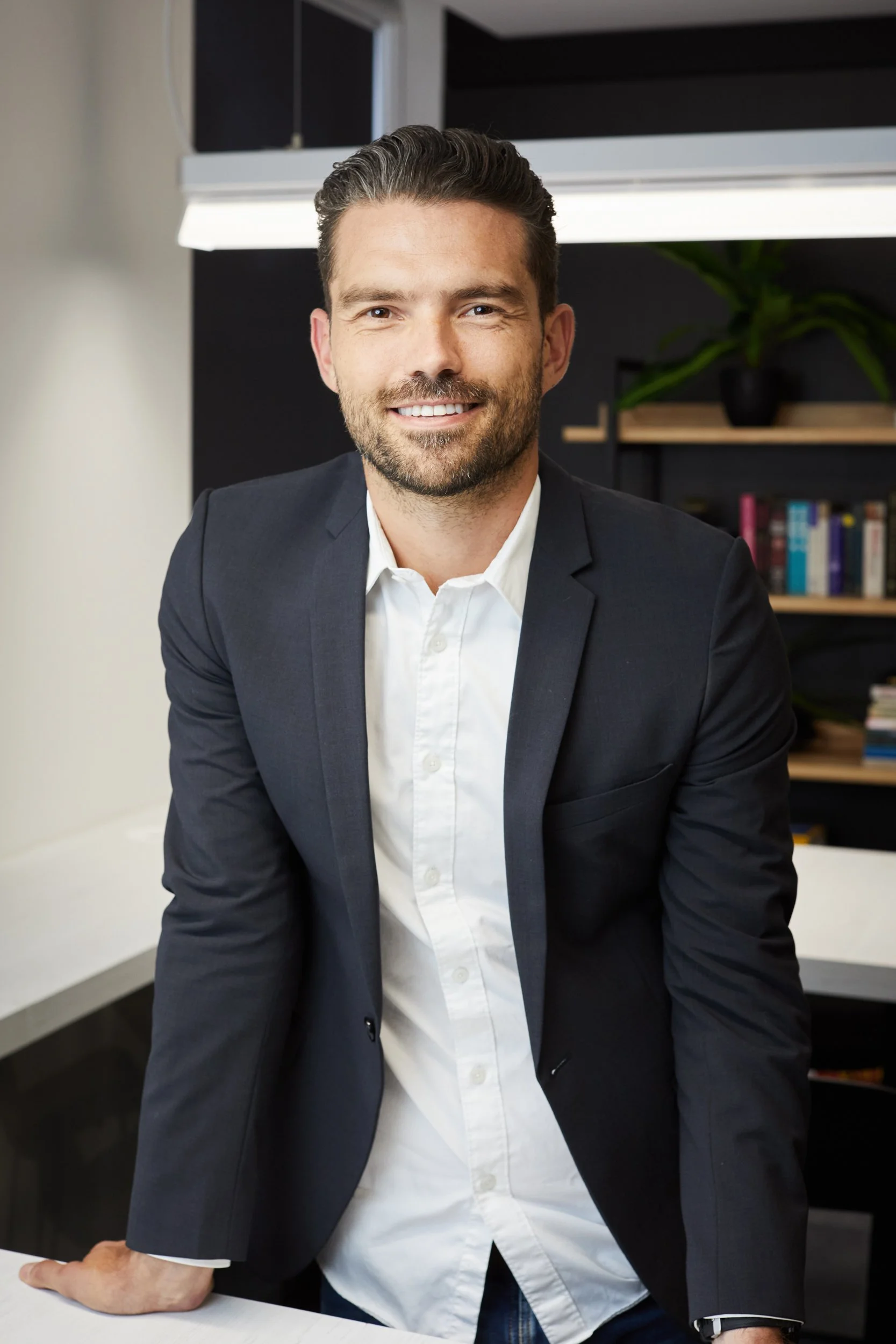 A smiling man in a black blazer and white shirt leaning on a white table in an office environment with a bookshelf and plant in the background.