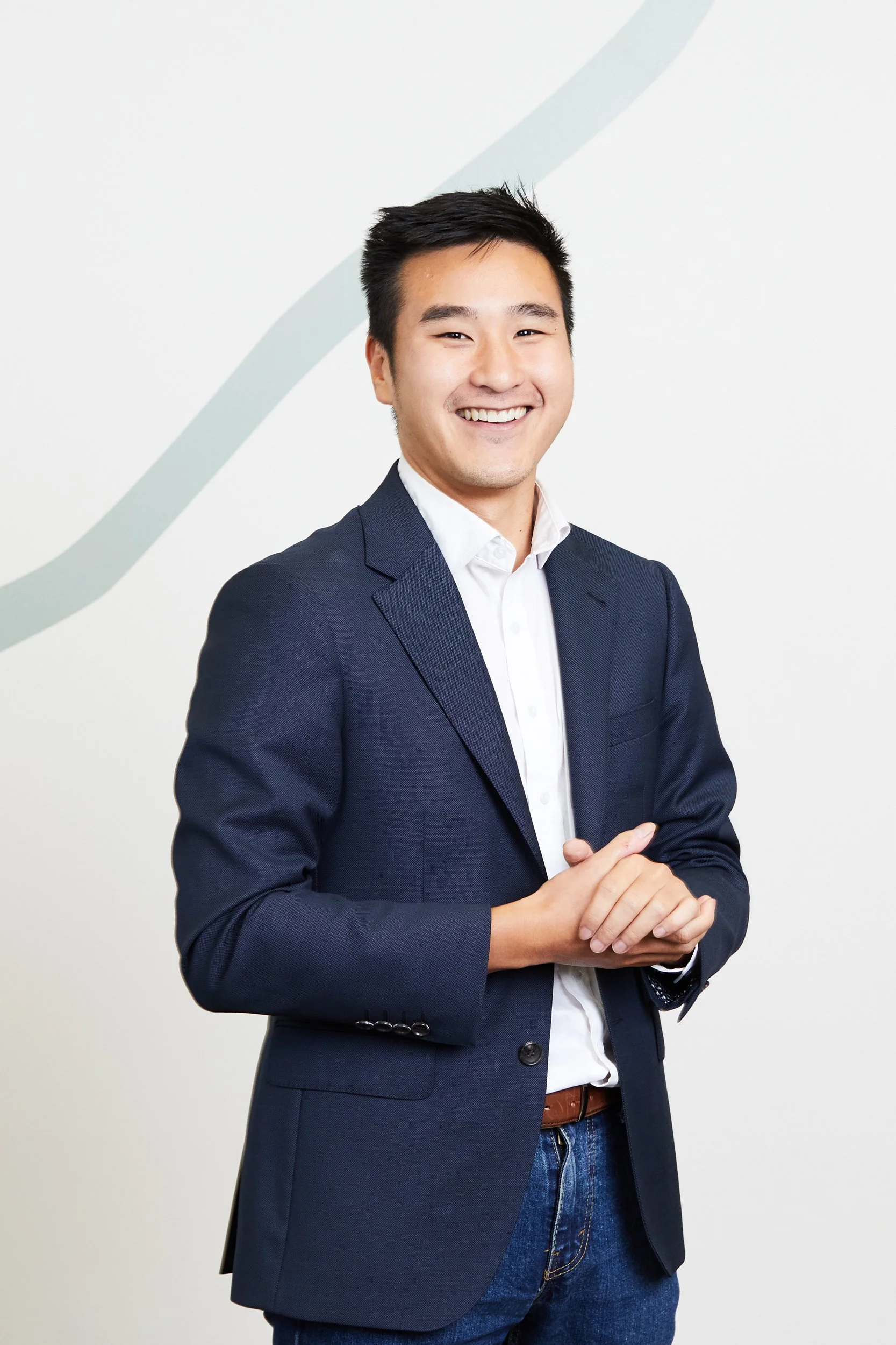 A young man in a navy blazer and white shirt smiling, standing against a light background with a subtle abstract line design.