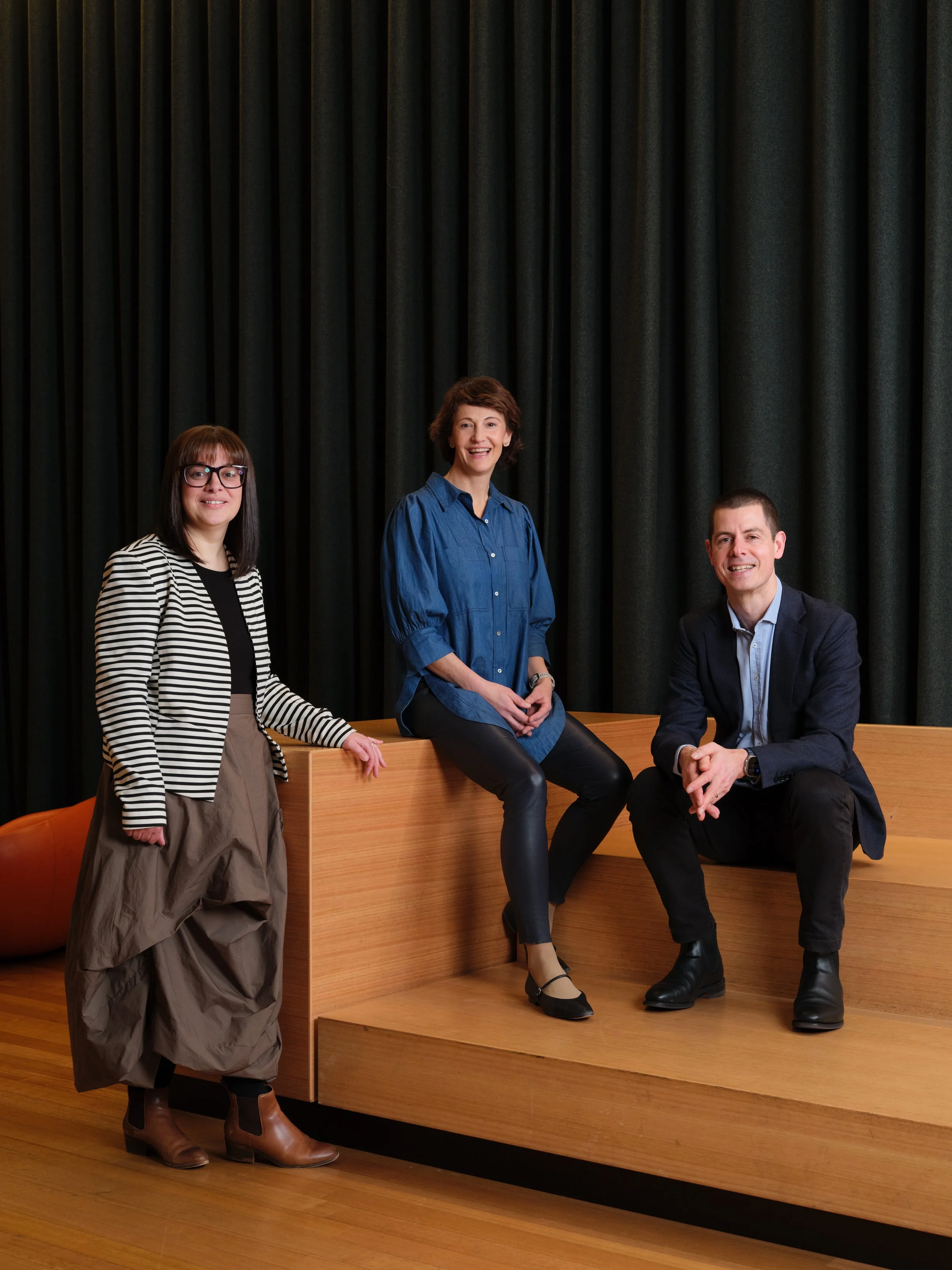 Three people posing in front of dark curtains, two women and one man, sitting and standing on a wooden platform.