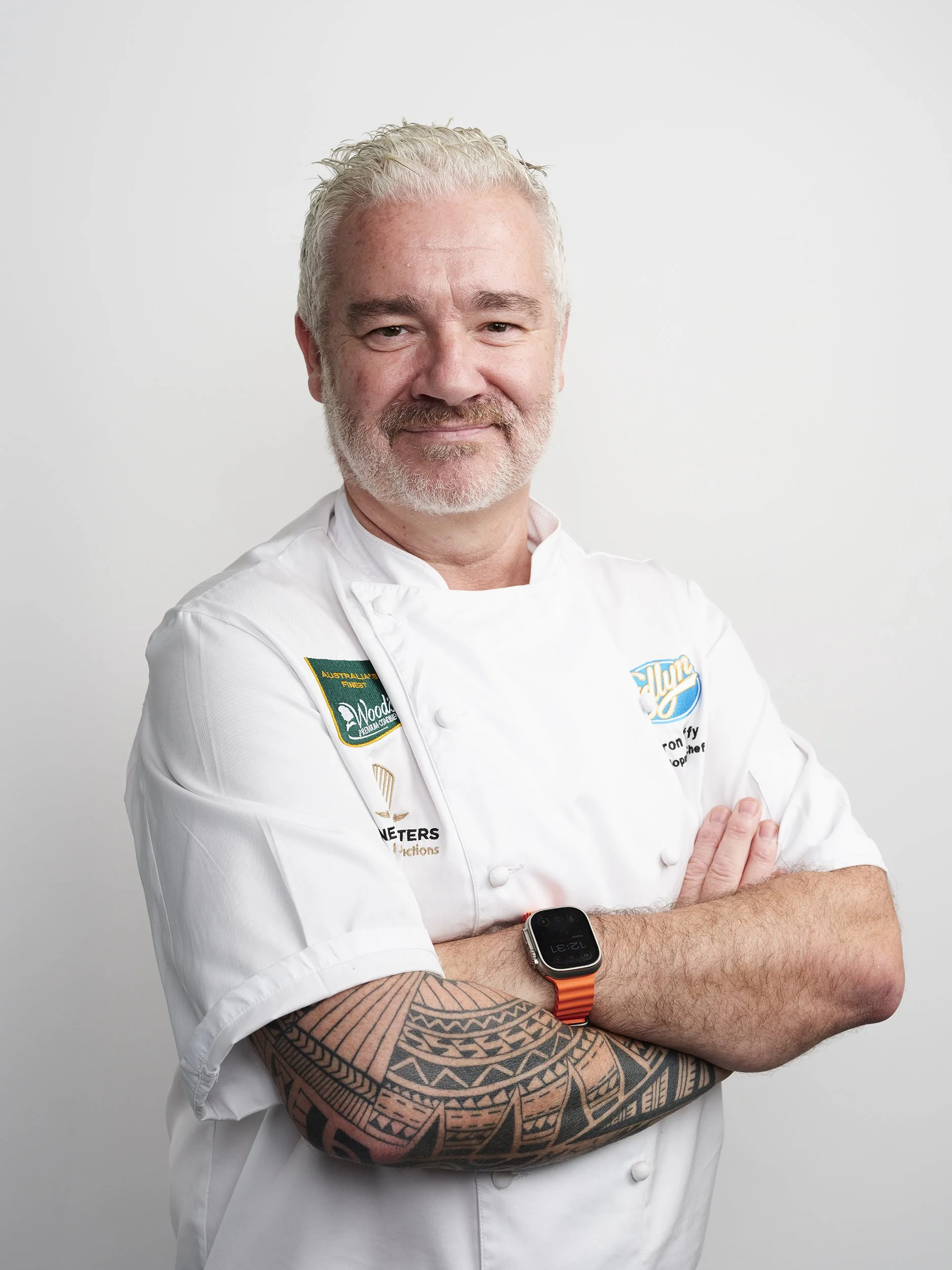 A male chef with gray hair and beard crossing his arms, wearing a white chef's jacket with various patches and an orange smartwatch, standing against a plain white background.
