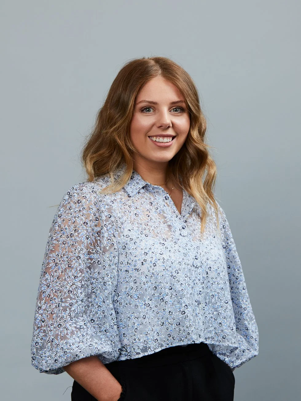 A young woman with shoulder-length wavy blonde hair smiling, wearing a light blue patterned blouse and black pants, standing against a plain gray background.
